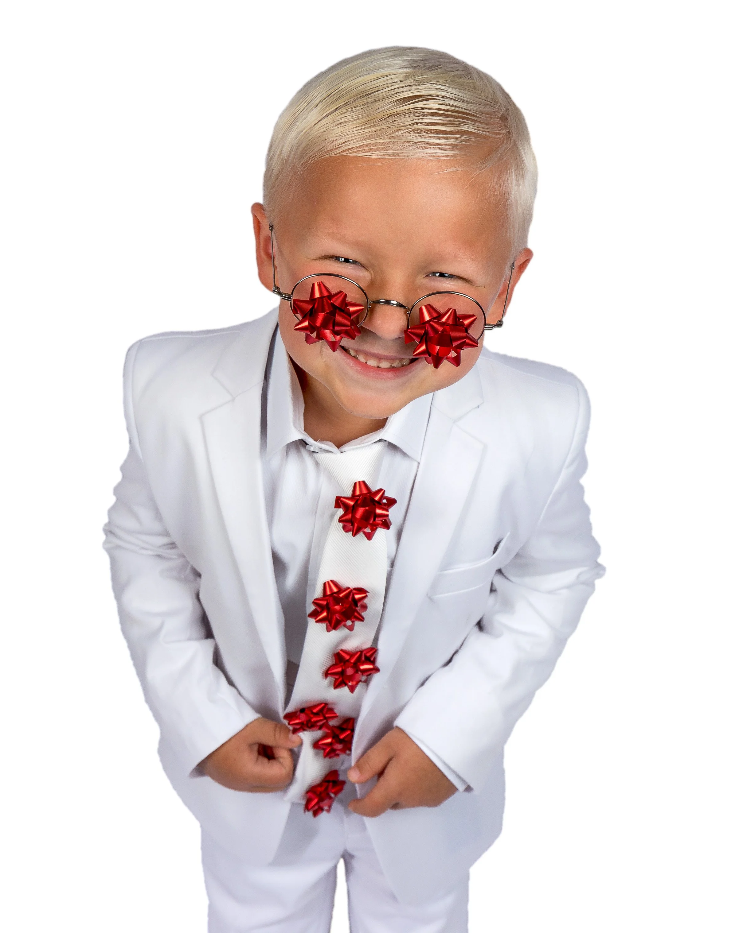 A young boy with blond hair and a big smile wearing a white suit and glasses decorated with red bows, with more red bows hanging down the front.