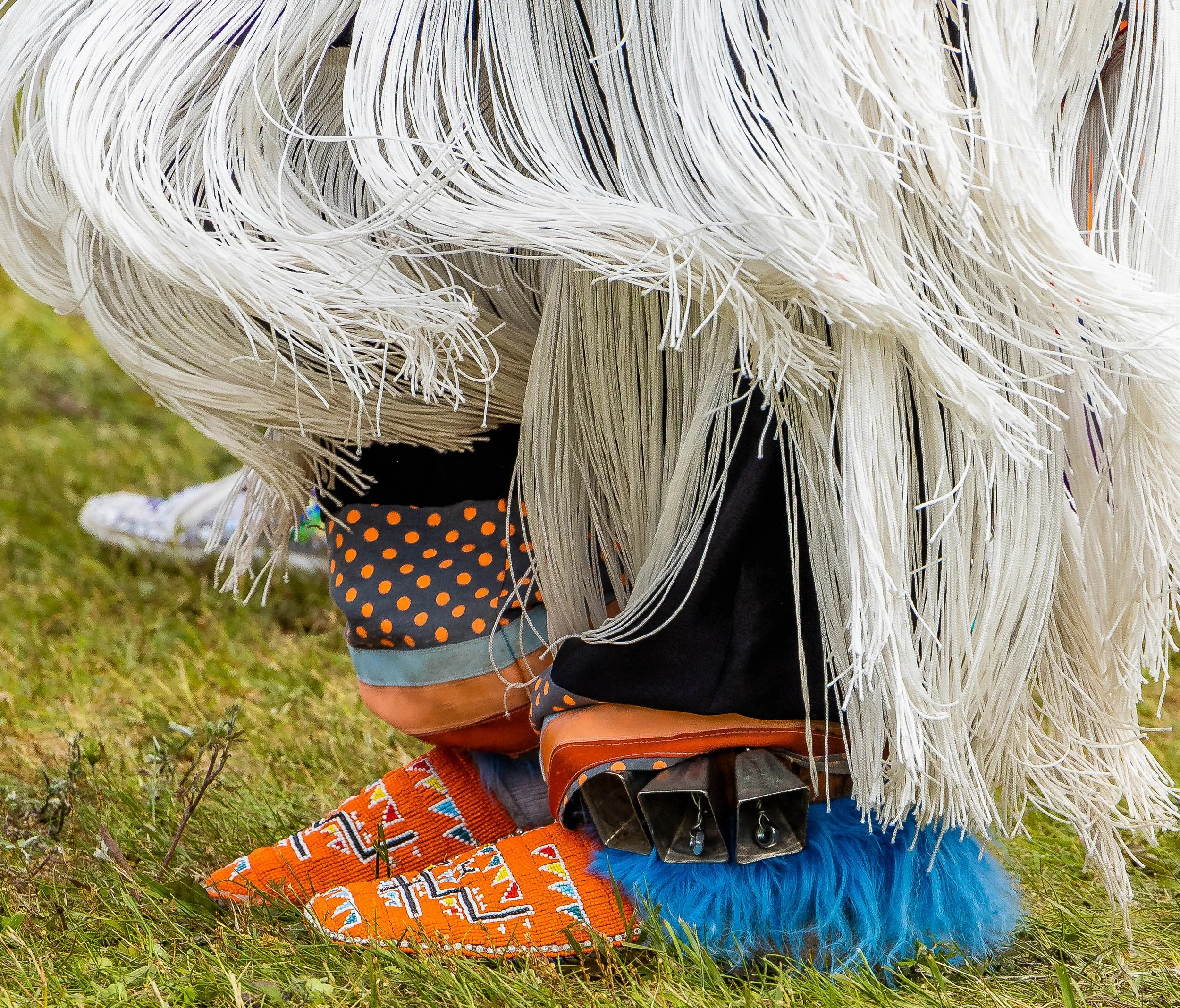 Close-up of a person wearing a traditional indigenous costume with a fringed headdress made of white fibers, colorful beaded accessories, and patterned clothing standing on grass.