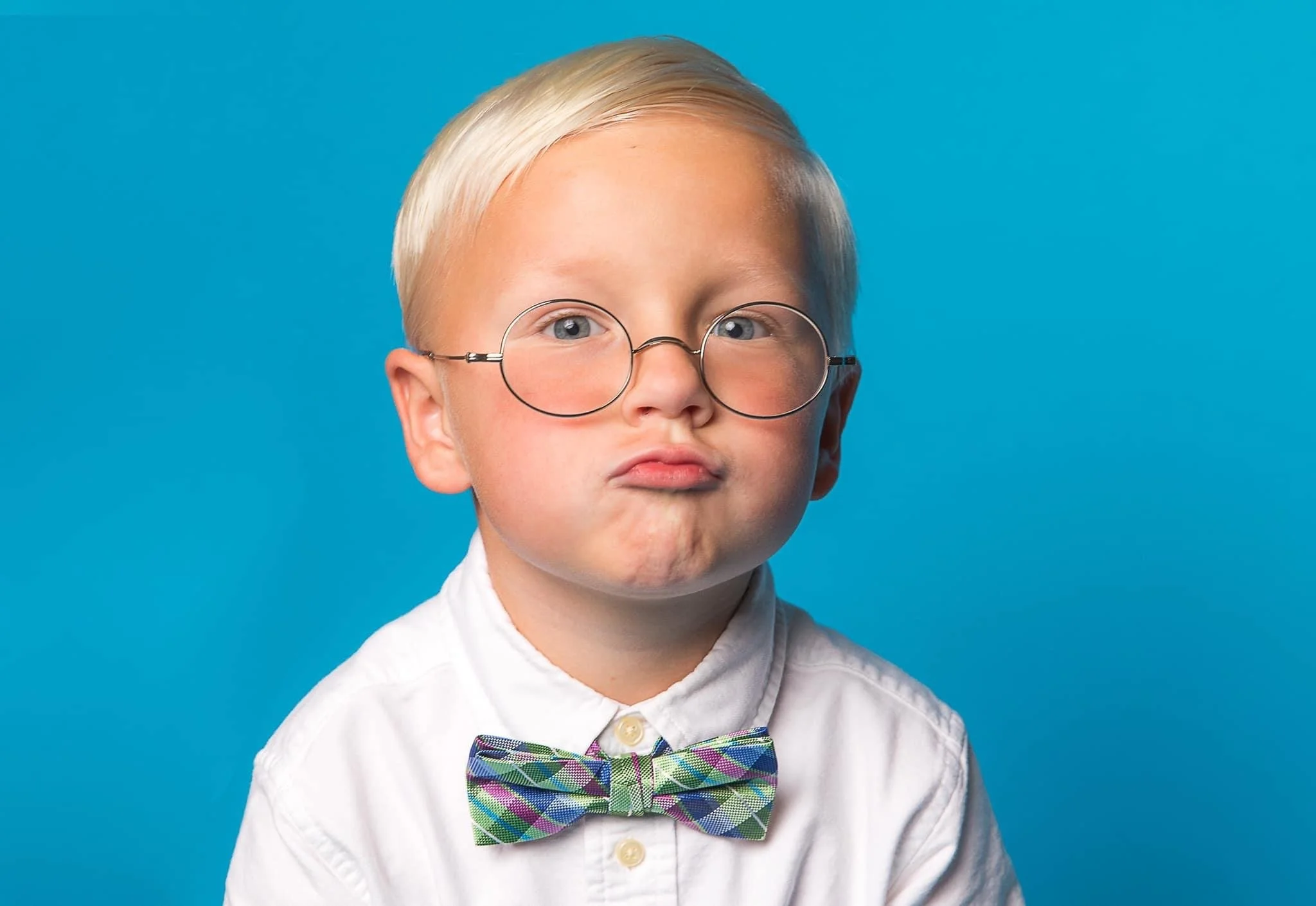 A young boy with blonde hair, wearing large round glasses, a white shirt, and a colorful plaid bow tie, stands against a blue background. His facial expression is pensive, with cheeks slightly puffed out and lips pressed together.