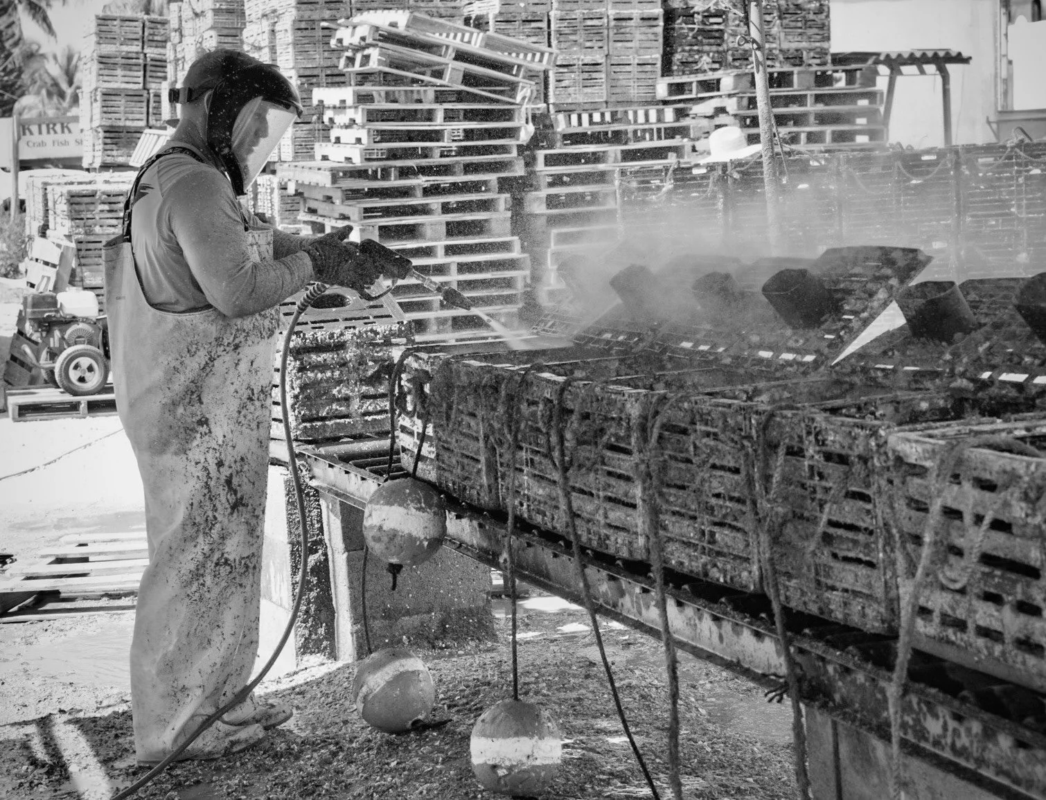 A worker wearing protective gear, including a helmet and apron, is using a power tool to cut large pieces of seafood on a processing table outdoors. There are stacks of crates and pallets in the background.