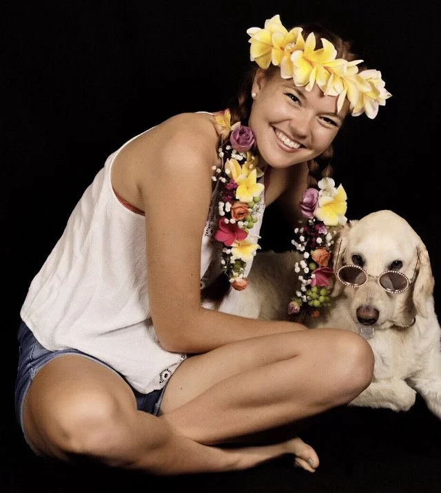 A young woman with long braided hair and light skin is smiling, wearing a flower crown and floral lei. She is sitting cross-legged on the floor with a golden retriever wearing sunglasses, both against a black background.