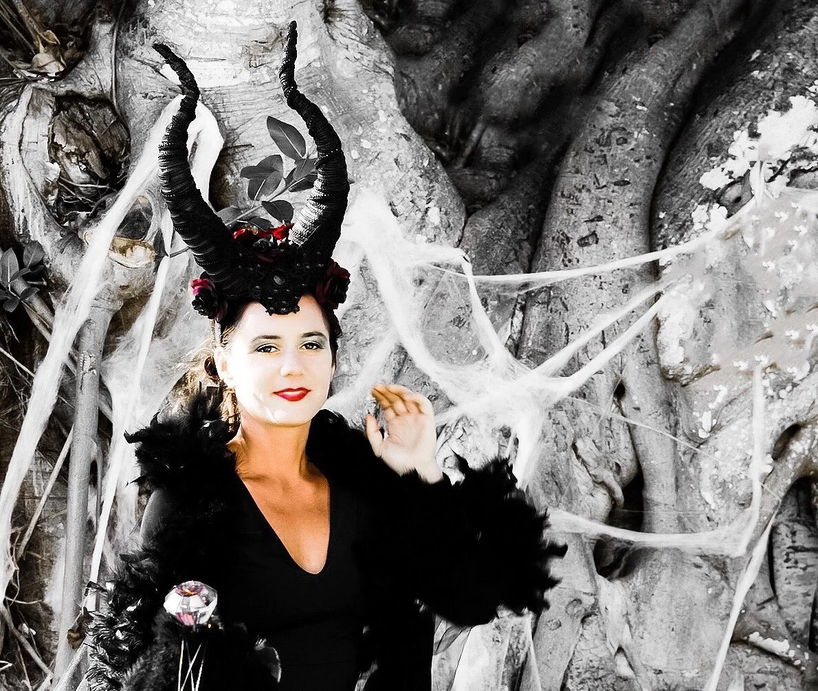 A woman dressed as Maleficent, wearing large black horns, black feathers, and a costume with feathers, standing in front of a large tree with cobwebs.