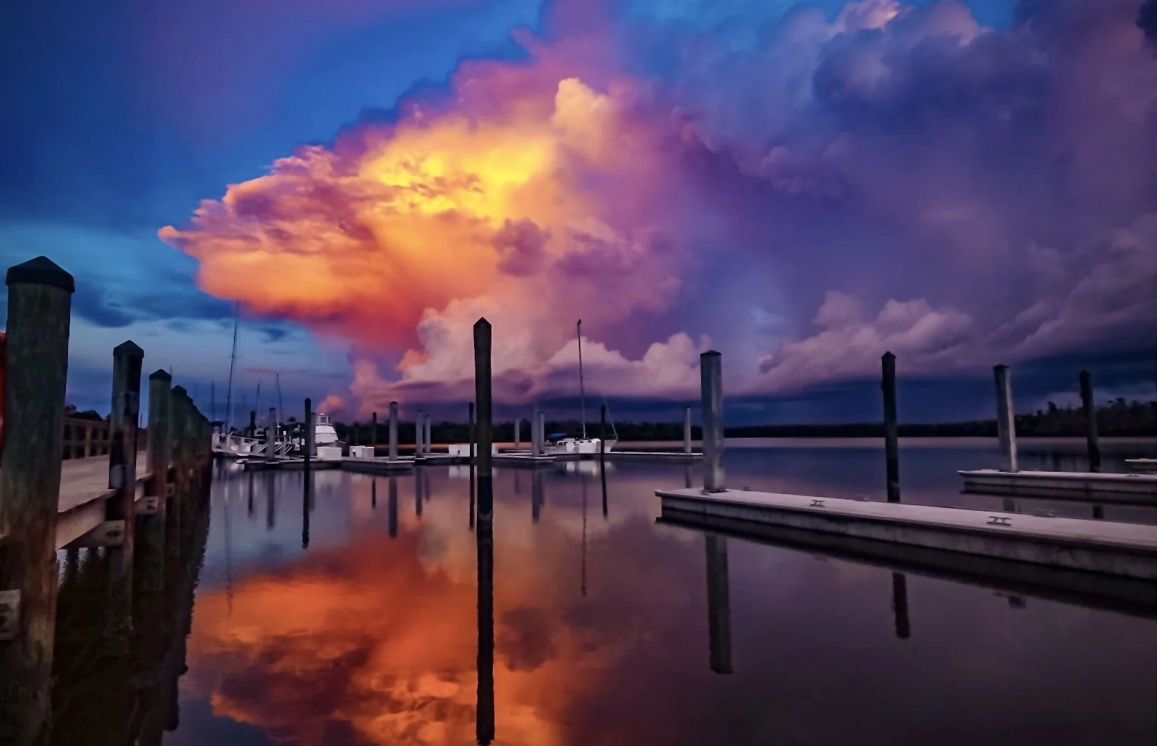 A marina at sunset with a vibrant, colorful sky reflecting on calm water, boat docks on either side, and docked boats visible in the background.