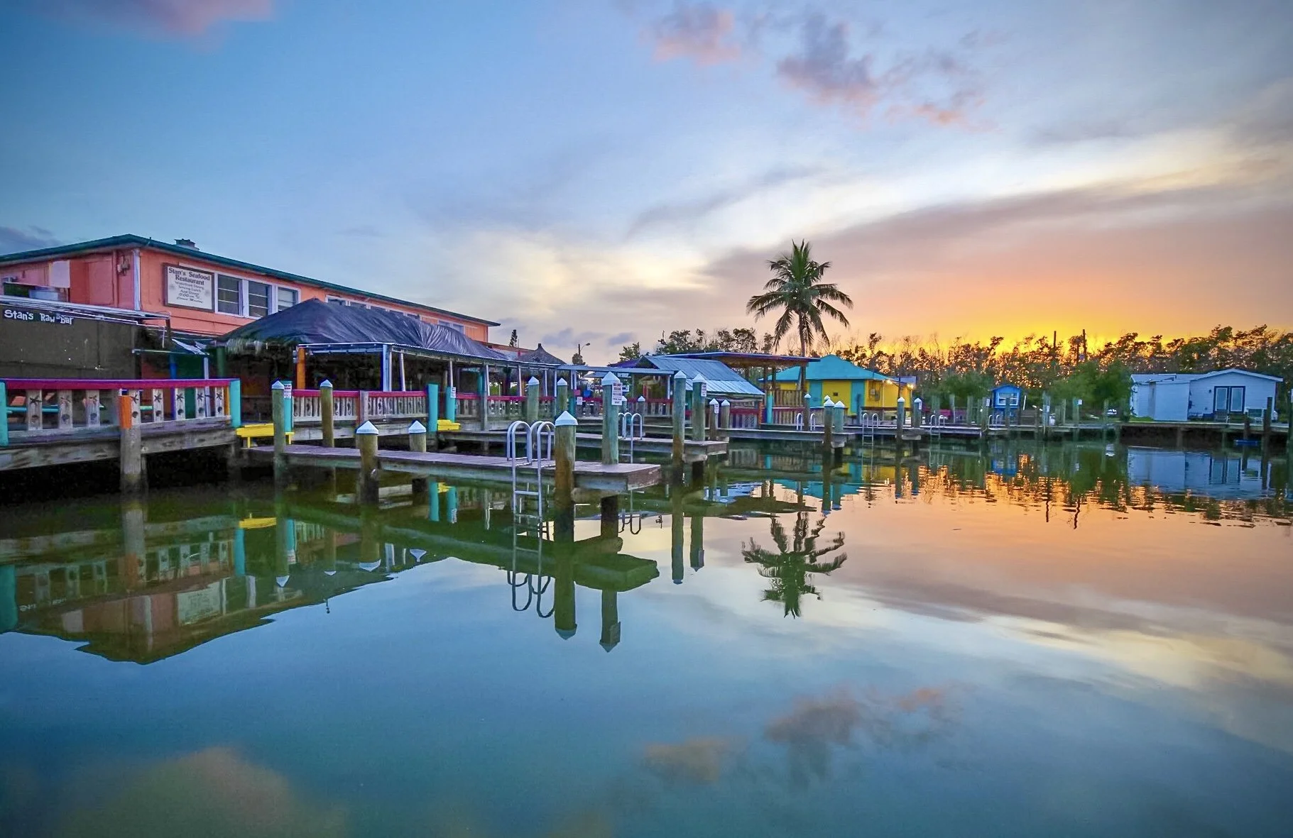 Colorful houses and a palm tree on a dock by calm water during sunset.