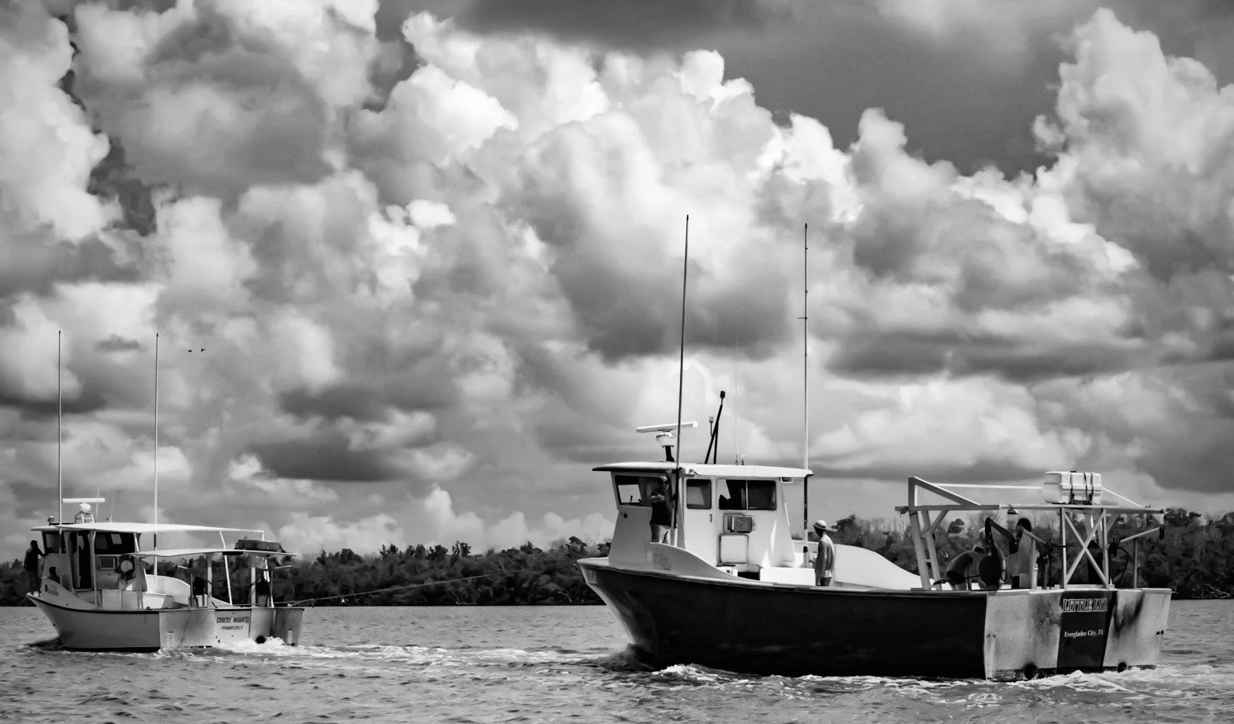Black and white photo of boats on the water with a cloudy sky in the background. One boat has a person standing on the deck, and there are antennas on some boats.