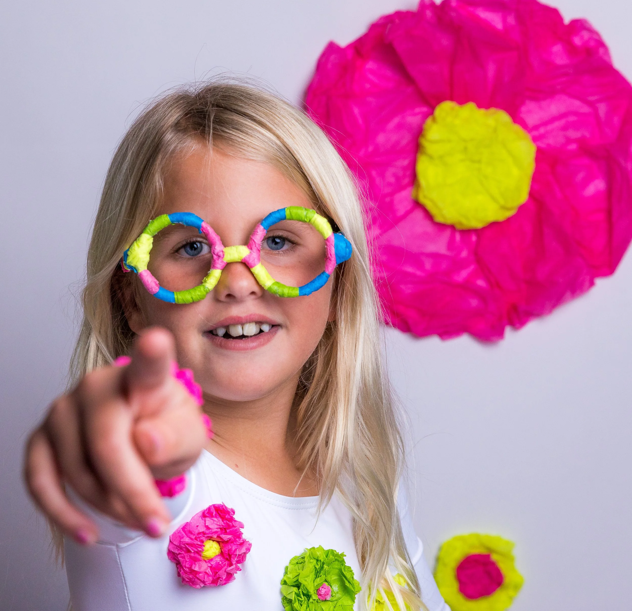 A young girl with blonde hair and blue eyes wearing colorful paper glasses and a white shirt decorated with paper flowers. She is smiling and pointing towards the camera, with a pink and yellow paper flower decoration in the background.