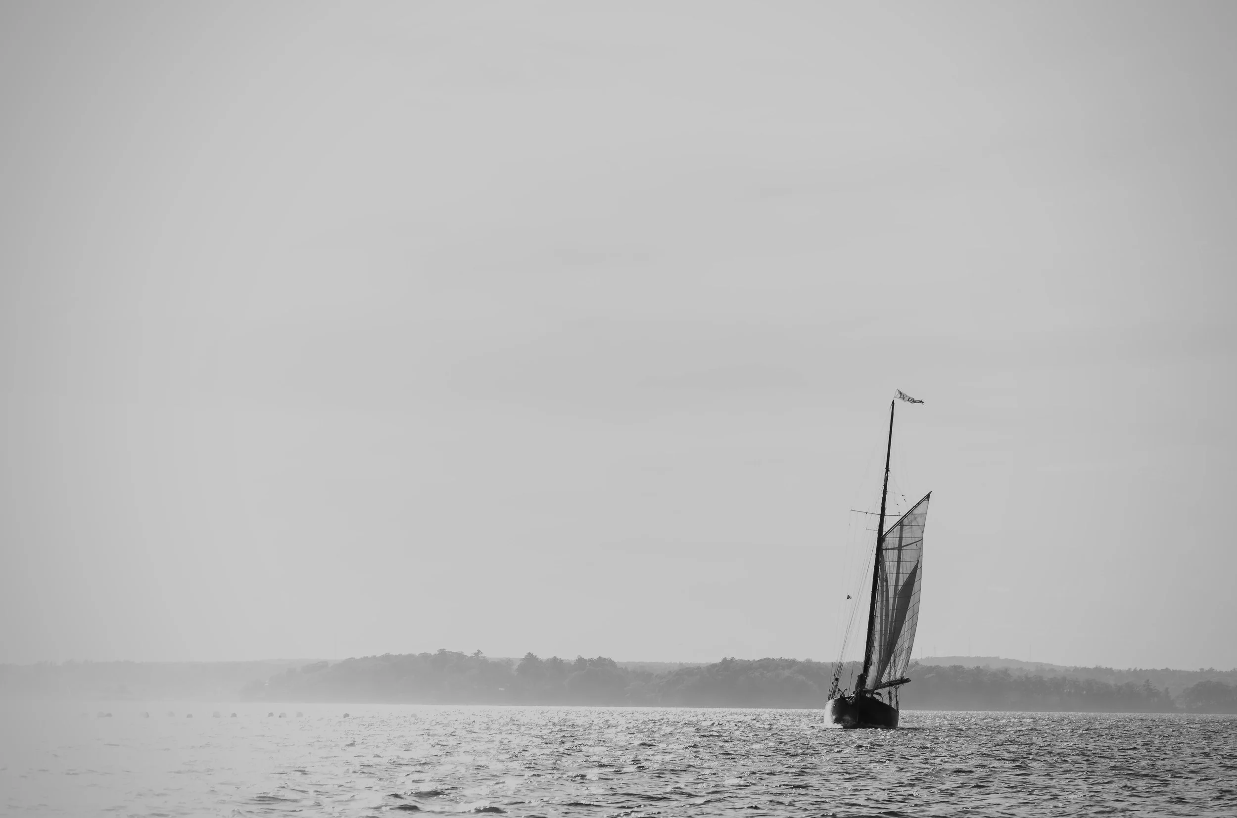 A sailboat with a large sail on a body of water, with a distant shoreline and cloudy sky.