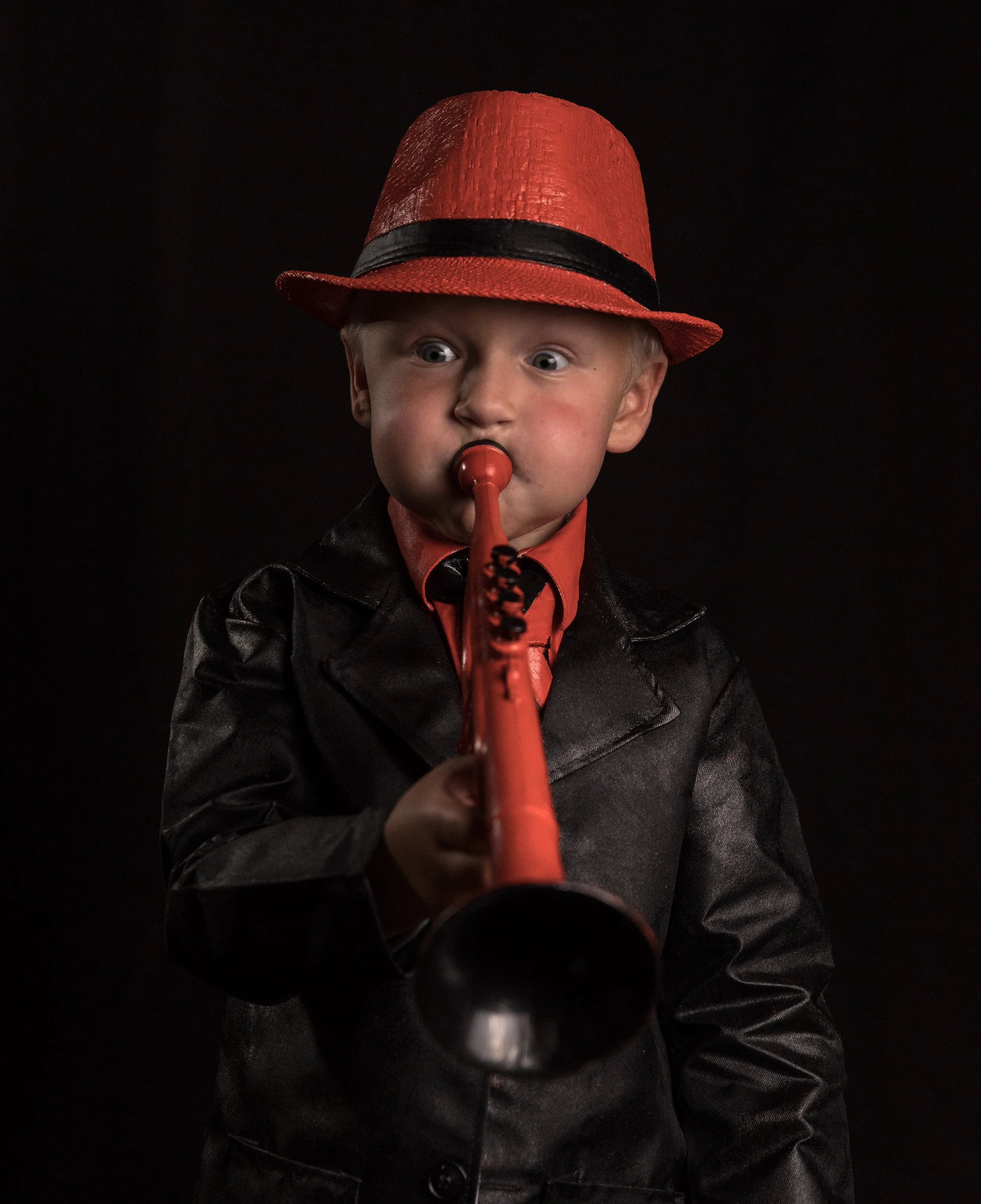 A young boy wearing a red hat with a black band, a black jacket, and a red shirt is playing a red recorder, with a black background.