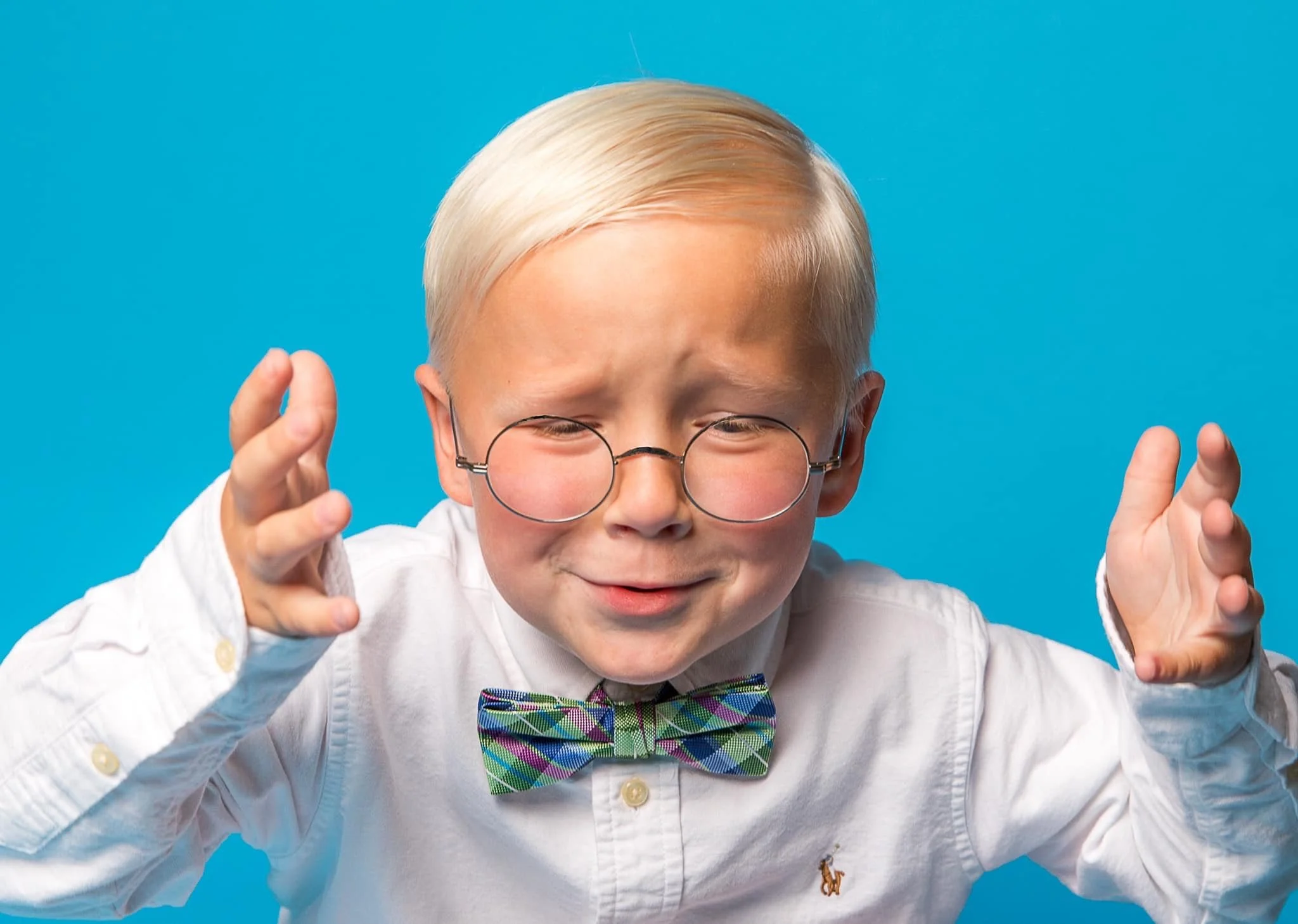 A young boy with blond hair and glasses wearing a white shirt and a colorful bow tie, standing against a blue background, making a gesture with his hands.