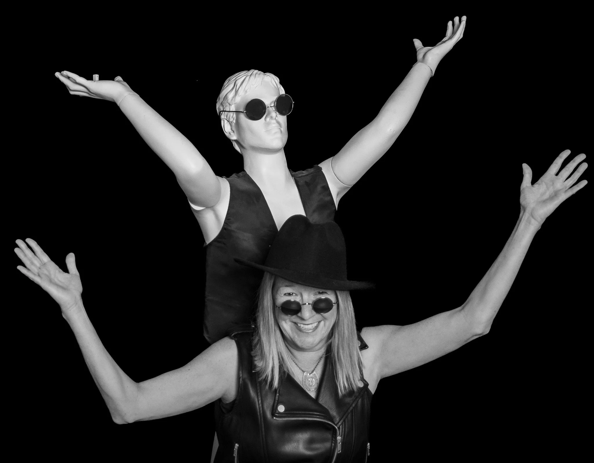 A woman with long hair, wearing sunglasses and a leather vest, smiling with arms raised, and a second person behind her with their arms raised up, both against a black background.