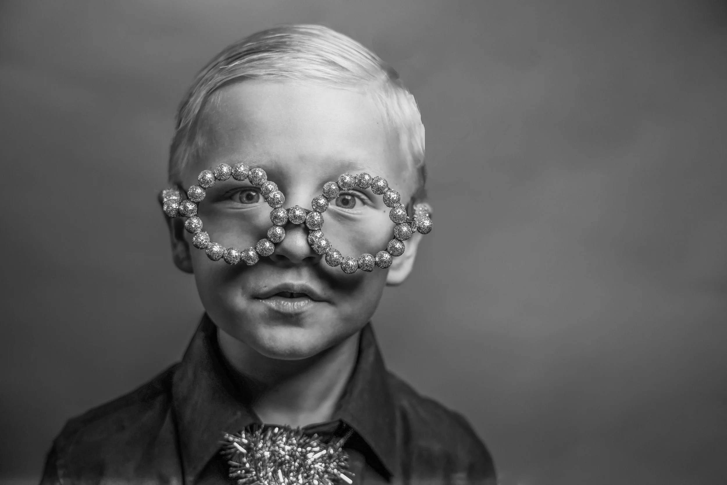 A black-and-white portrait of a young person with short, slicked-back hair, wearing novelty glasses made of glittery beads, and a jacket with a large floral brooch on the lapel.