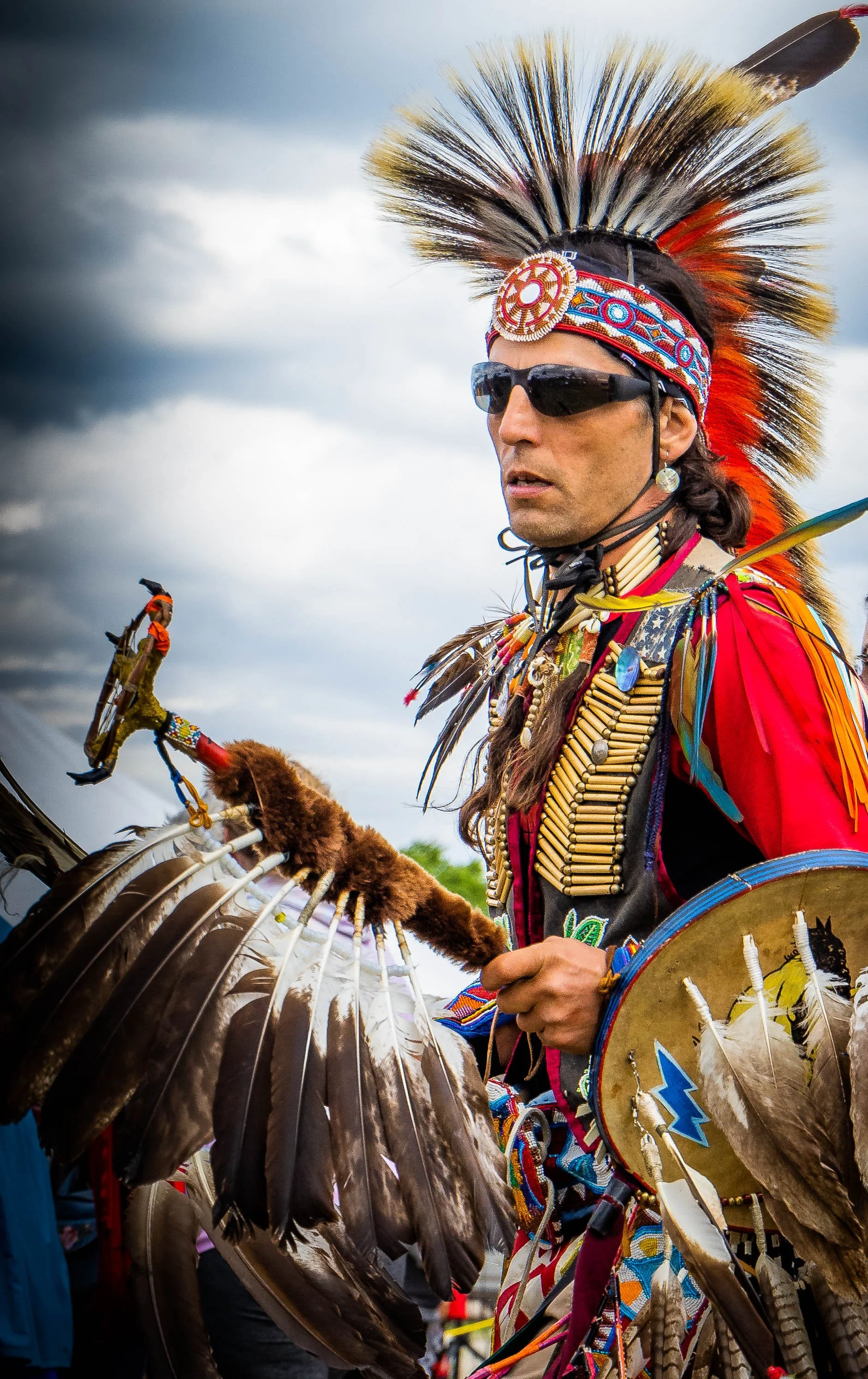 A Native American man wearing traditional headdress, sunglasses, and beaded accessories, holding a shield and a staff with feathers, outdoors with cloudy sky.