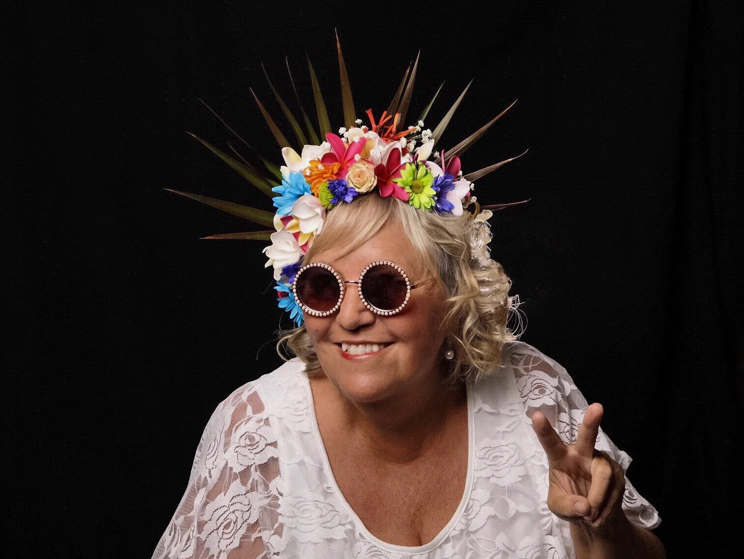 A smiling woman wearing pink sunglasses decorated with rhinestones, a floral crown with colorful flowers and greenery, a white lace top, and making a peace sign with her hand against a black background.