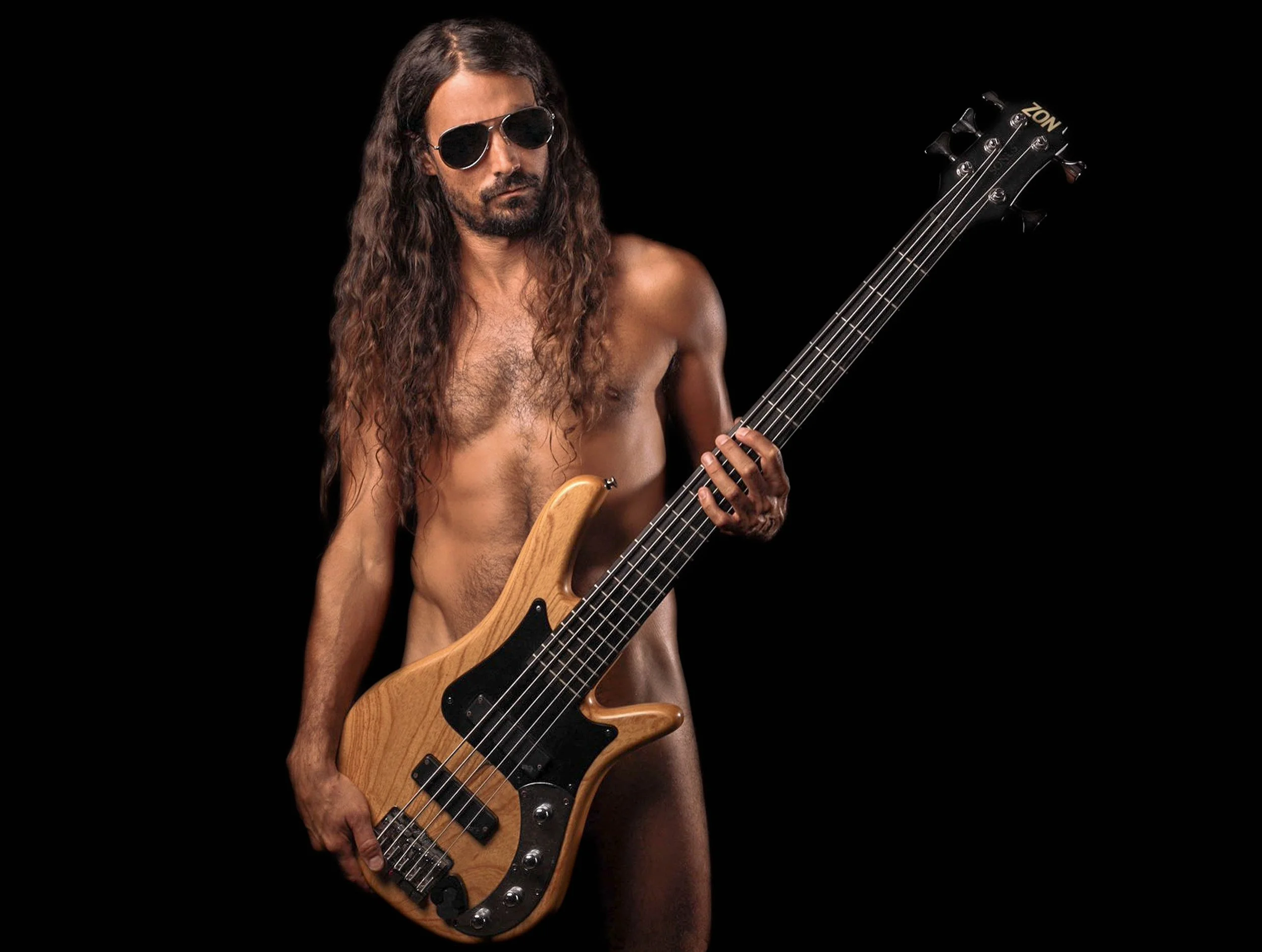 A shirtless man with long hair and sunglasses holding a bass guitar against a black background.