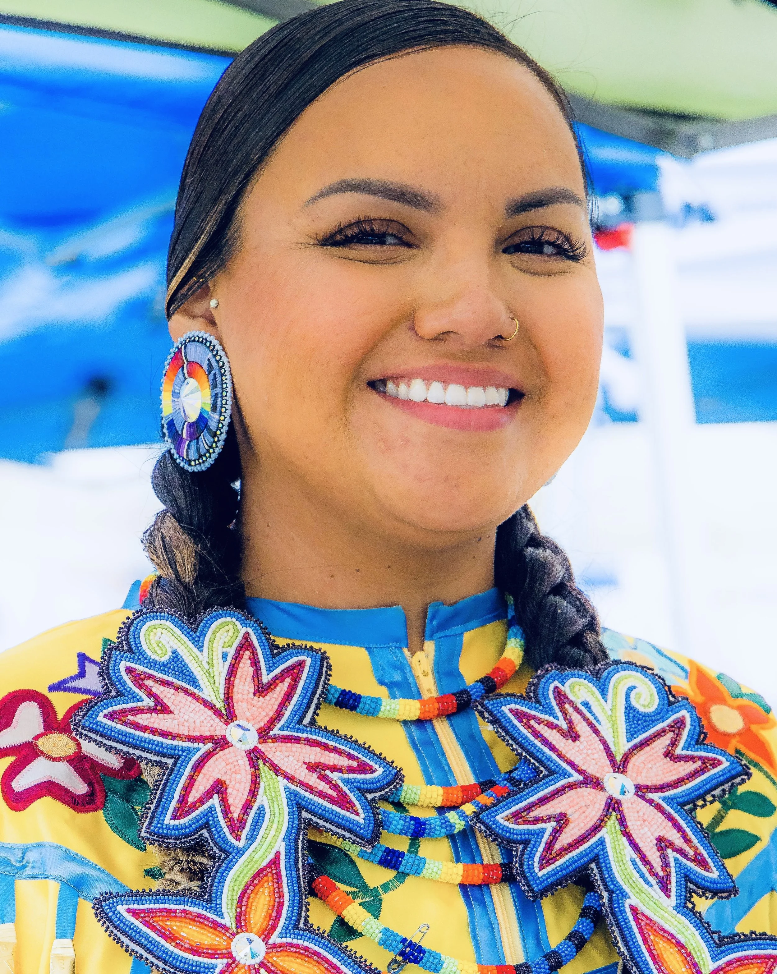 Close-up of a woman wearing traditional colorful beadwork clothing and jewelry, smiling with dark hair in braids, and a nose ring, standing outdoors.