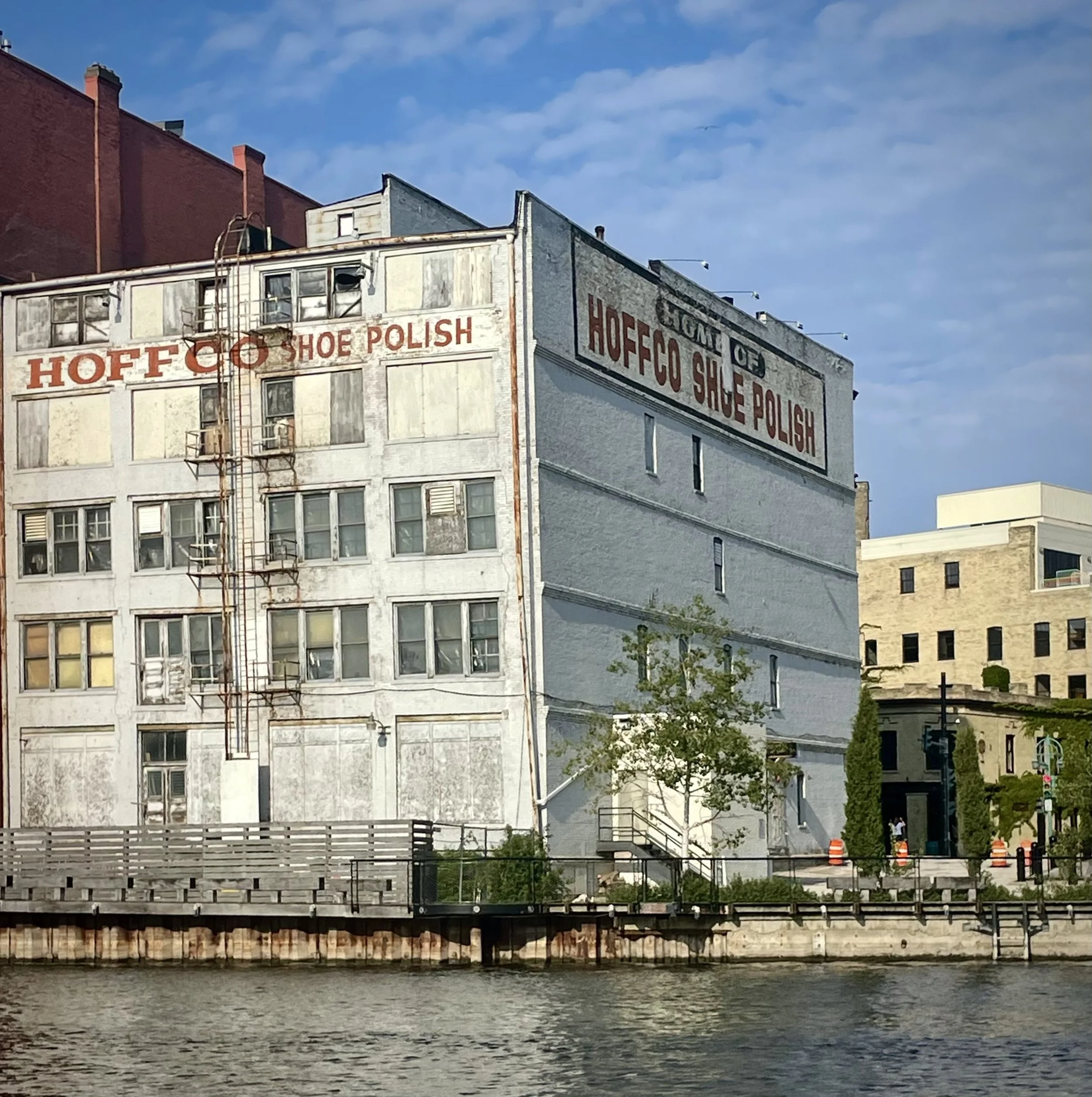 View of a river with buildings along the shoreline, including a white building with red lettering that reads 'HOFFCO SHOE POLISH', and other structures with fire escape ladders and signage under a blue sky.