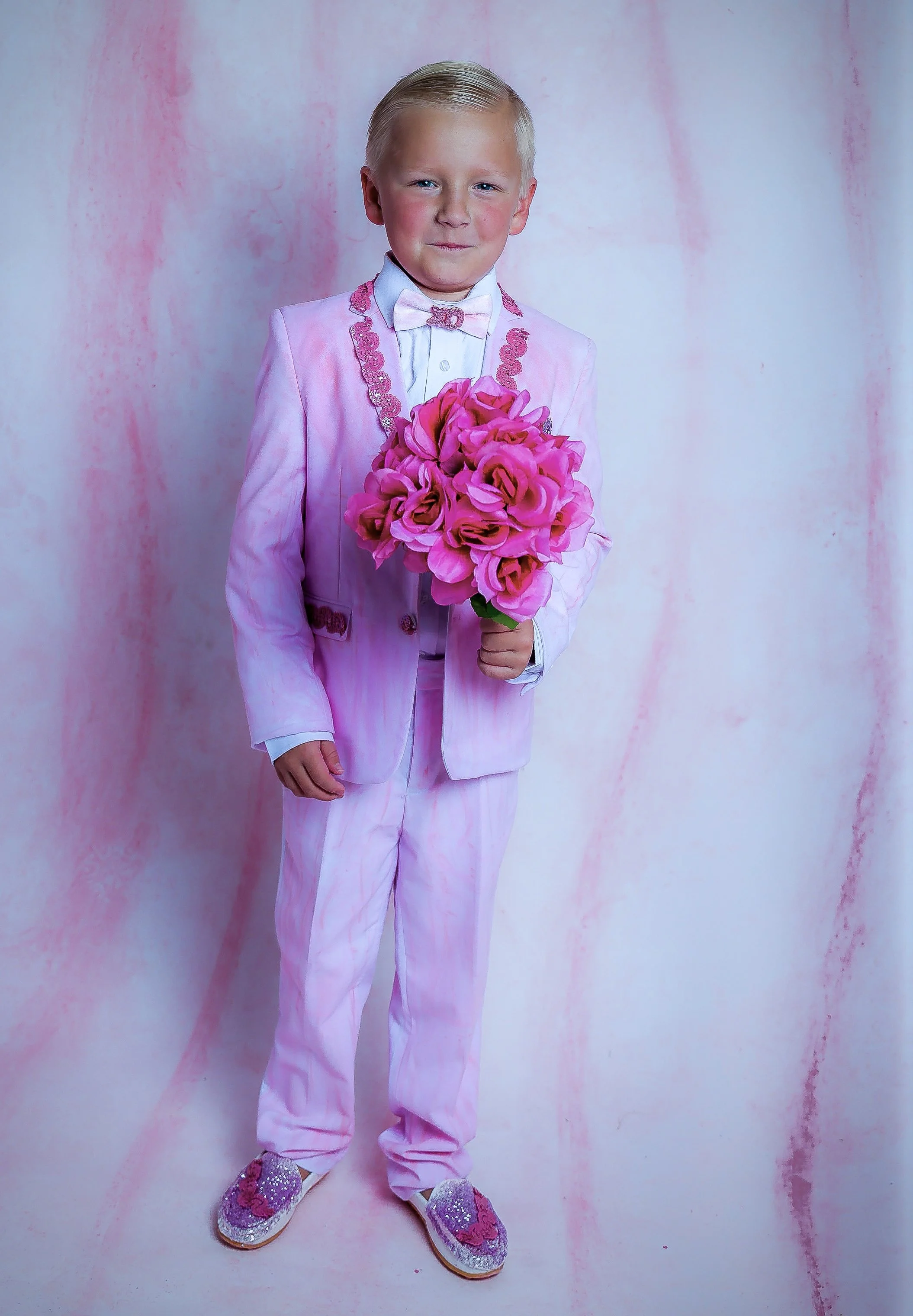 A young boy dressed in a pink suit and bow tie, holding a bouquet of pink flowers, standing against a pink and blue background.