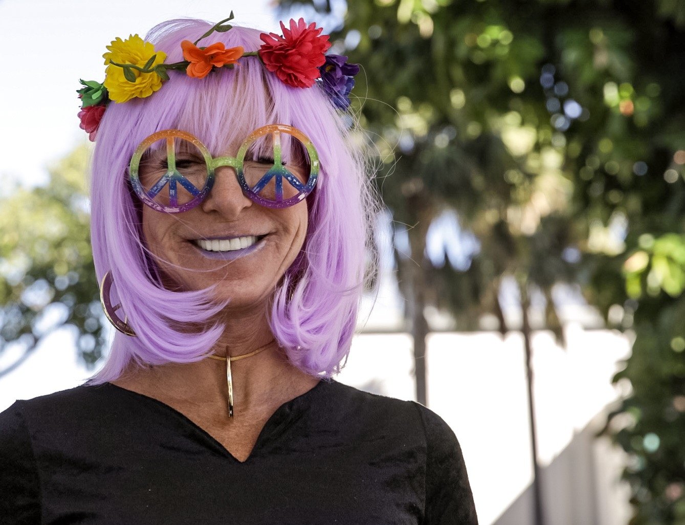 Woman with lavender hair wearing rainbow floral crown, large peace sign glasses, and black top outdoors.