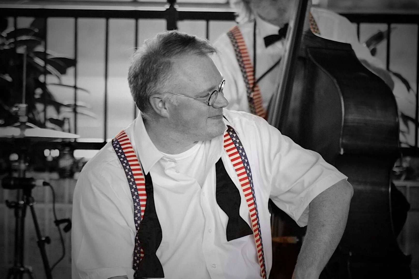 A man wearing glasses, a white shirt, and suspenders with an American flag pattern is smiling and playing a double bass in a black-and-white setting.