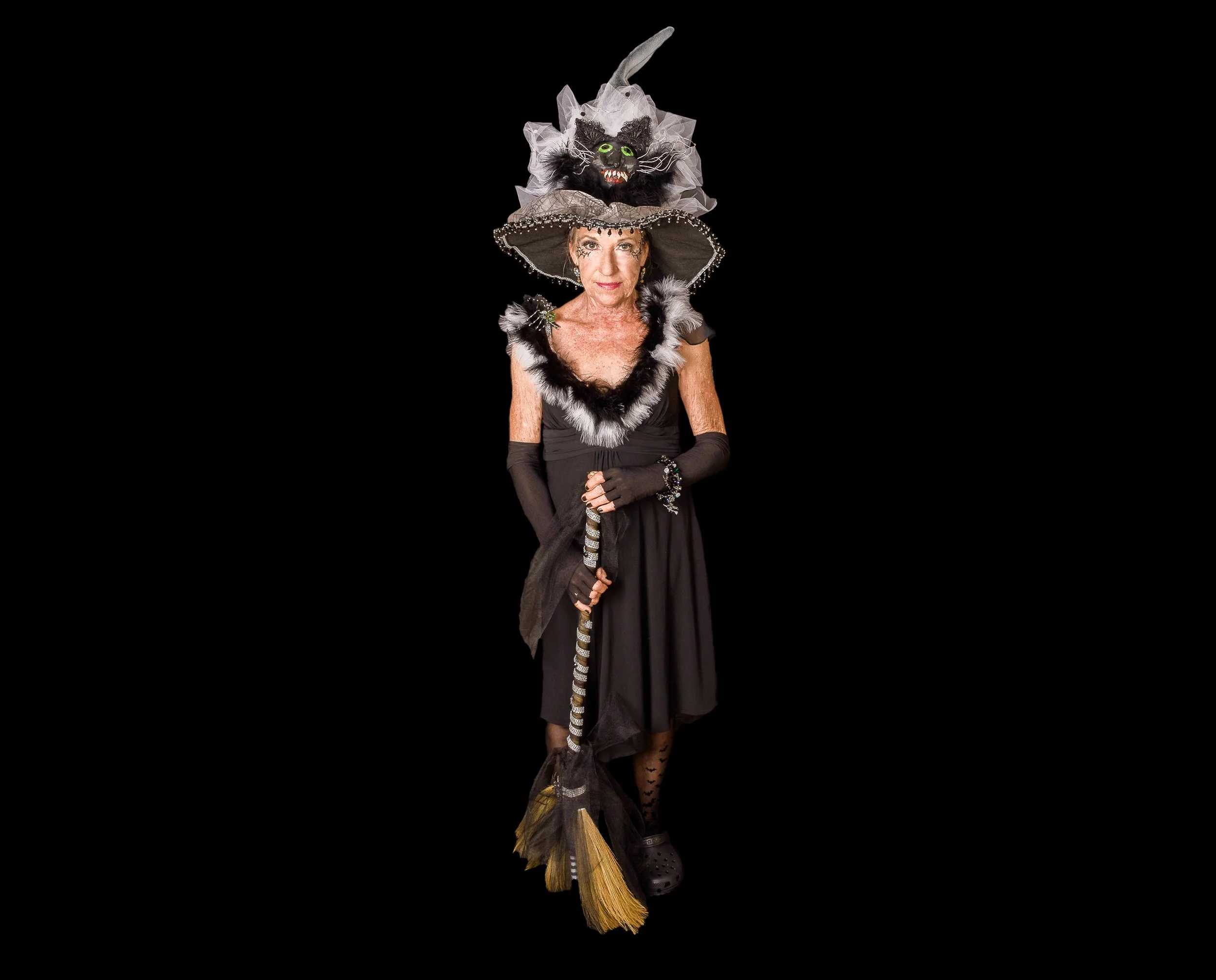 Woman dressed as a witch with a black dress, long gloves, and black fishnet stockings, wearing a large witch hat with a black cat face on top, holding a broom, against a black background.