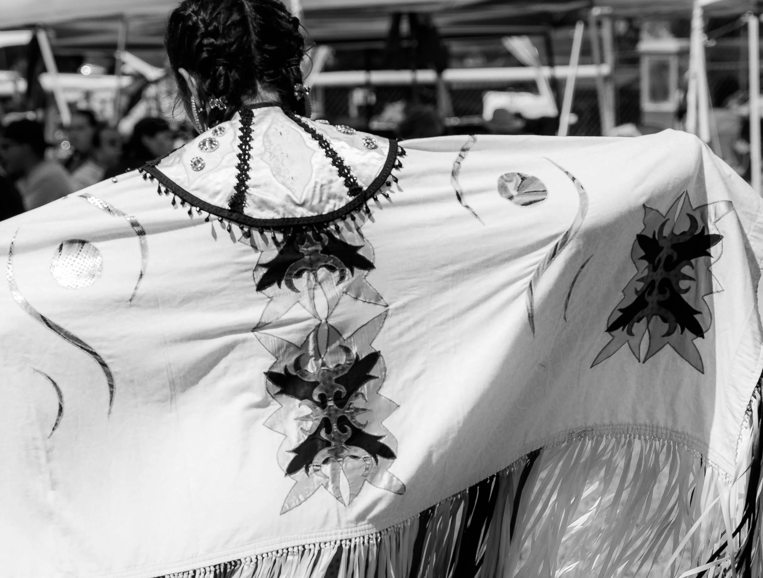 The back of an individual with short, curly hair, wearing earrings and a decorative top, standing behind a table with a patterned tablecloth with fringes, in a busy outdoor market or festival setting.