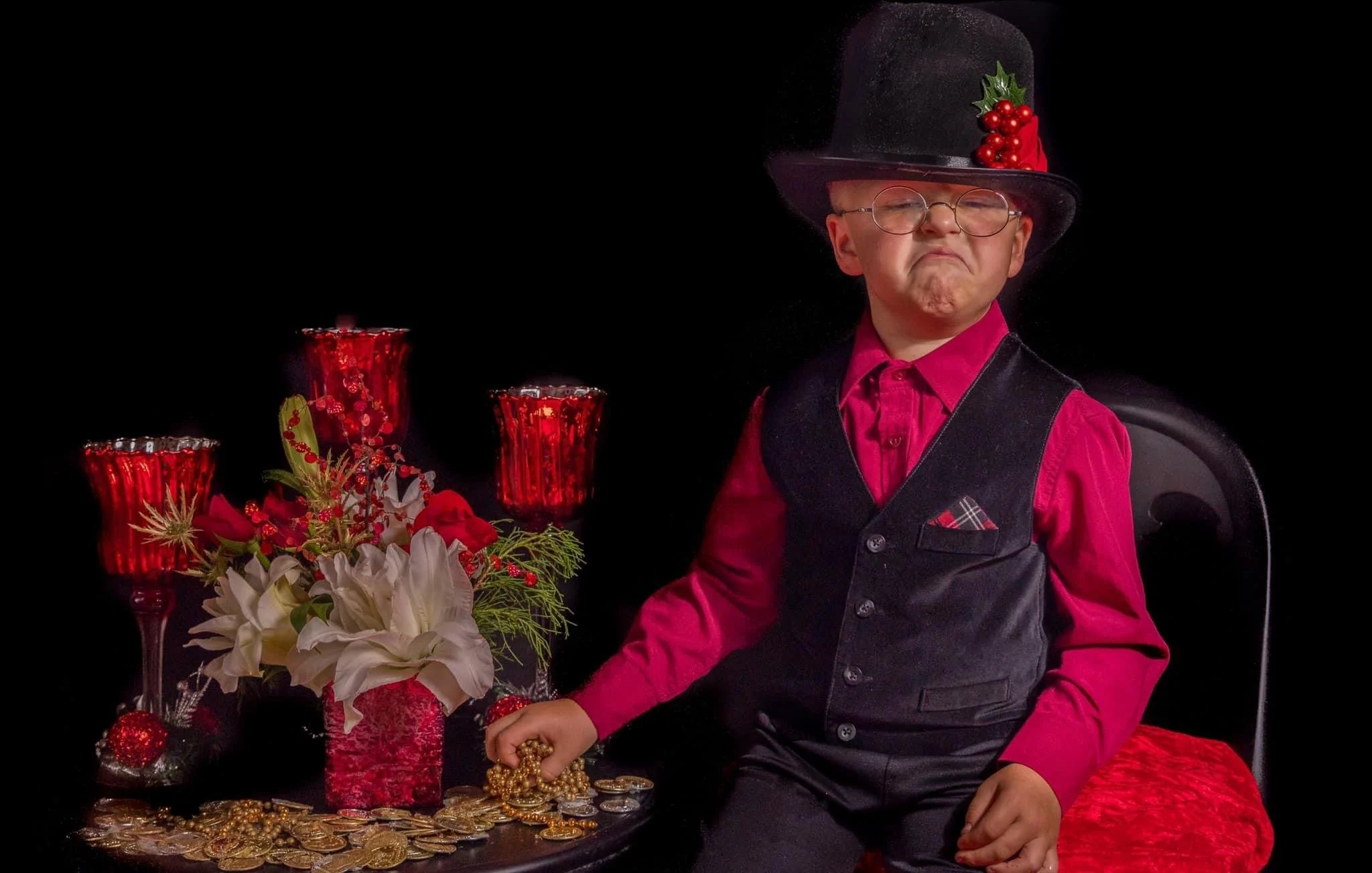 A young boy wearing glasses, a red shirt, and a black vest sits next to a table filled with red and gold decorations, including candles, flowers, and coins, styled for Christmas.