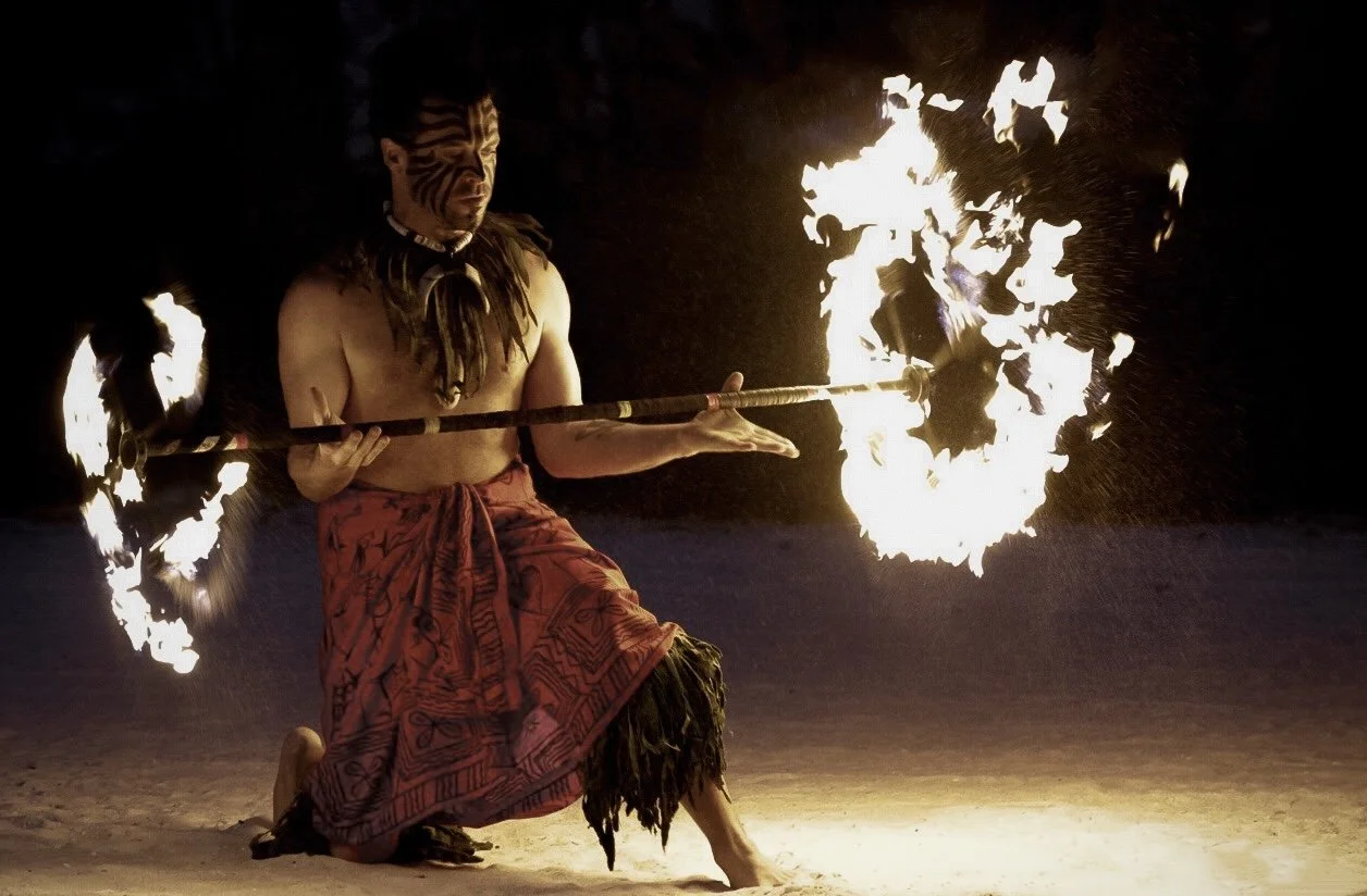 A man dressed in traditional Polynesian attire performs a fire dance at night, holding a stick with flames on both ends, with flames illuminating the dark background.