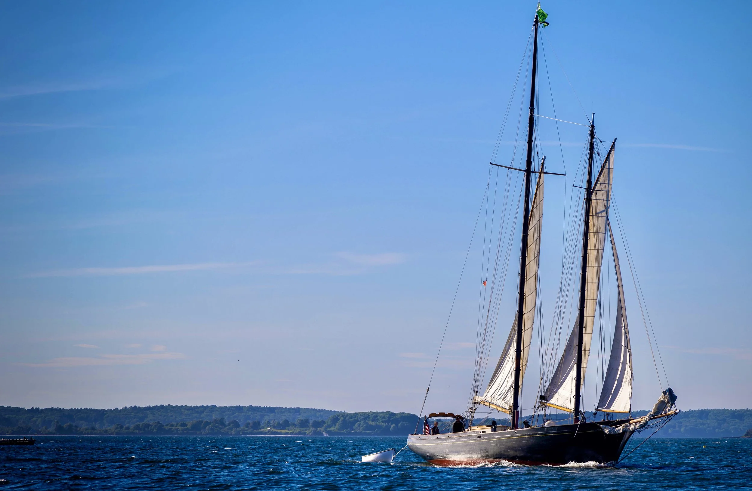A large sailboat with white sails on a body of water during daytime, with a distant shoreline and a clear blue sky.