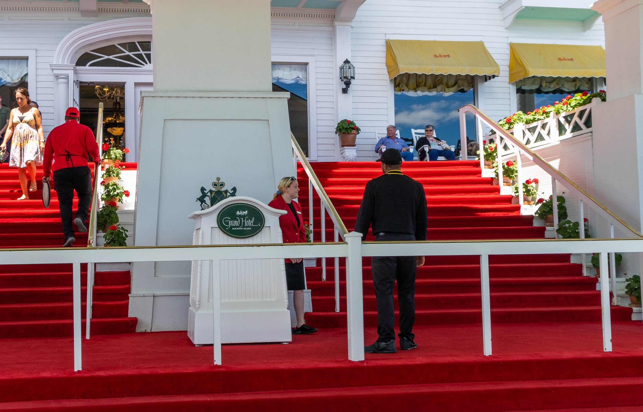 People walking up a red carpeted staircase outside a white building with yellow awnings, gabled windows, and decorative flower pots. A woman in a colorful dress and a man in red attire are descending the stairs, while a woman in a red jacket and a ma