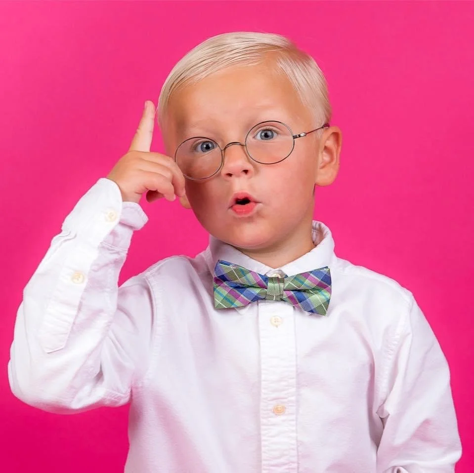 A young boy with blonde hair and glasses adjusting his glasses against a pink background. He is wearing a white shirt and a colorful plaid bow tie, with a surprised expression.