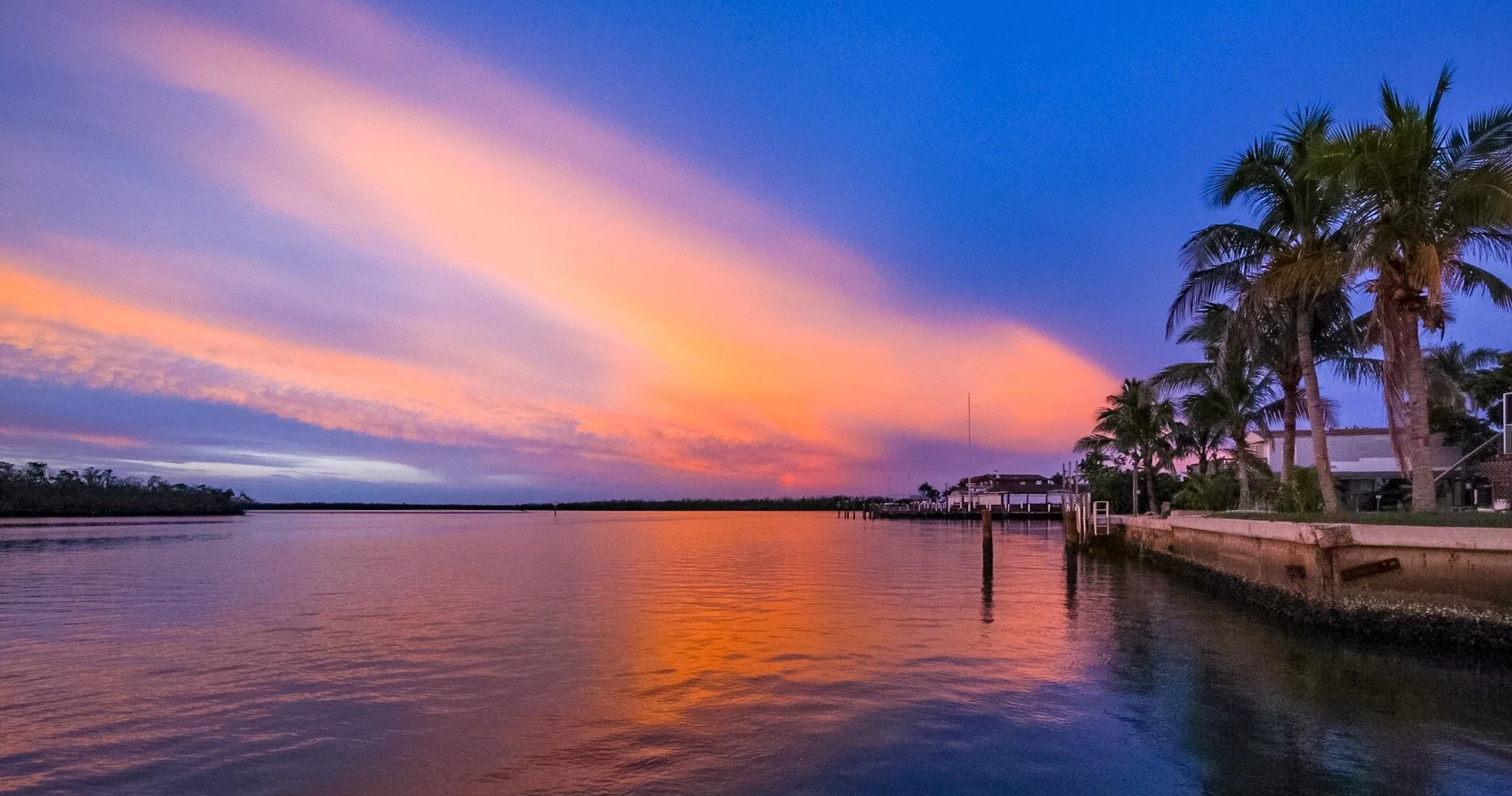 A colorful sunset over a calm river, with pink, purple, and orange hues in the sky, palm trees along the bank, and a dock extending into the water.