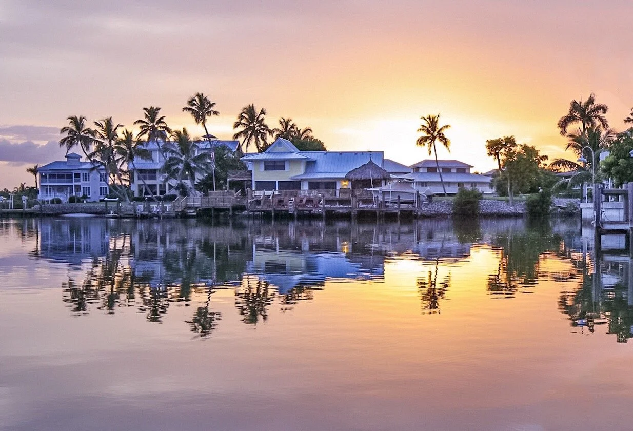 Sunset over a waterfront with houses and palm trees, reflecting in the calm water.