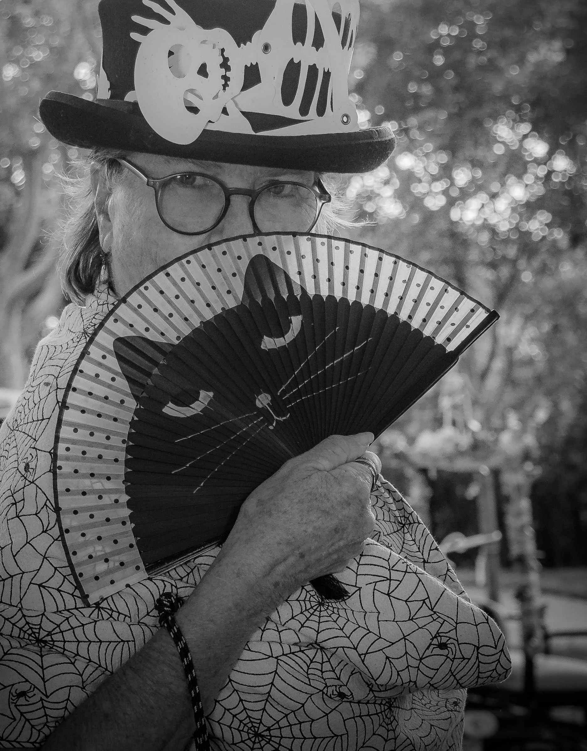 An elderly person wearing glasses and a patterned shirt holds a decorative fan in front of their face. They are also wearing a hat with a skeleton design, featuring a skull and ribs. The background is blurred with bokeh effect.