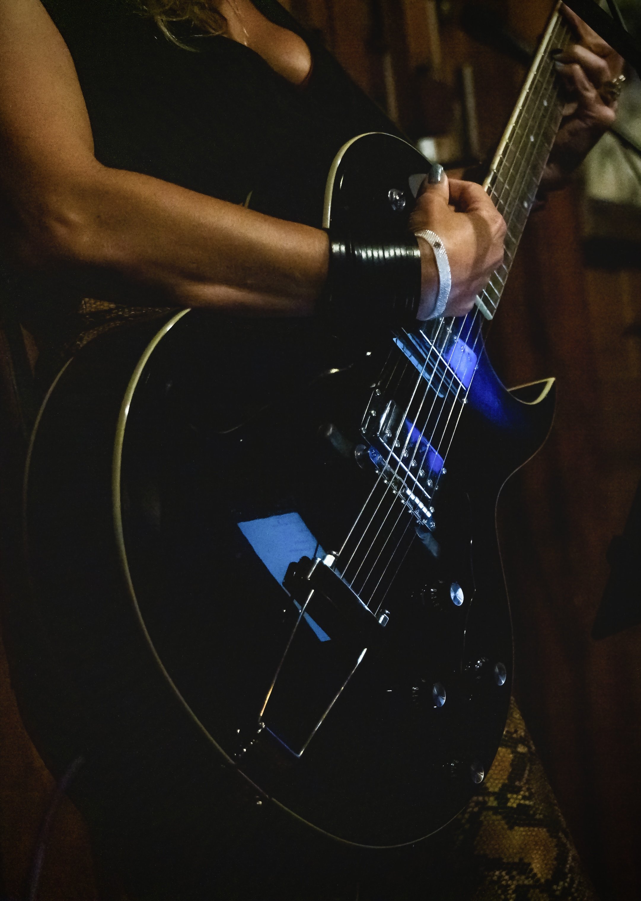 Close-up of a person playing an electric guitar in a dimly lit room. The individual is wearing a black outfit and silver accessories, with a focus on their hand strumming the guitar strings.