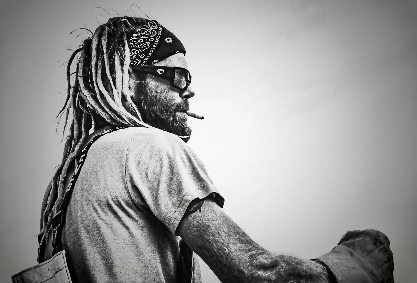 Black and white photo of a man with dreadlocks, wearing sunglasses, bandana, and a t-shirt, smoking a cigarette, with an arm extended outward.