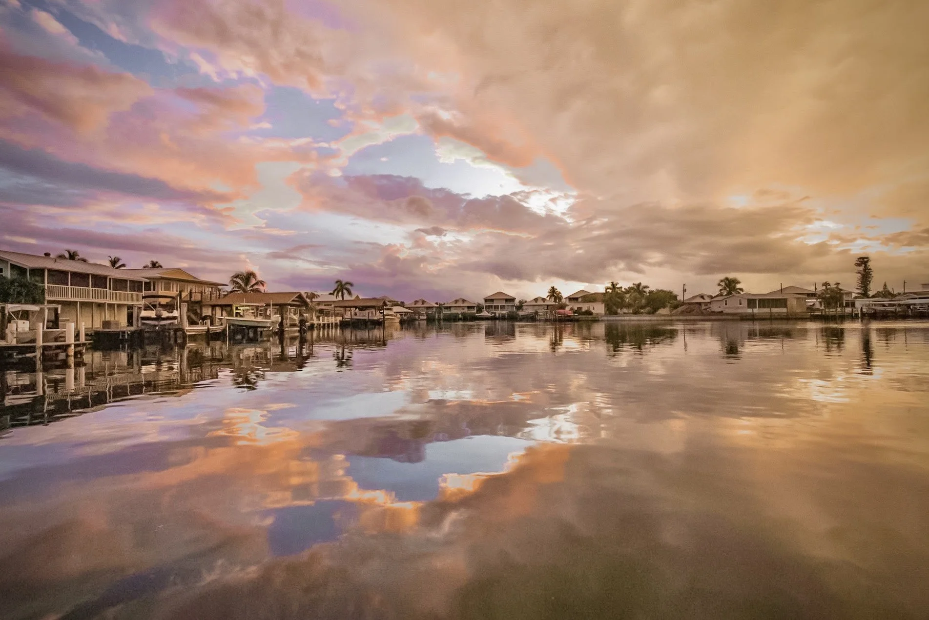 Sunset over a calm waterfront neighborhood with houses, palm trees, and boats, reflecting vibrant pink and orange clouds in the water.