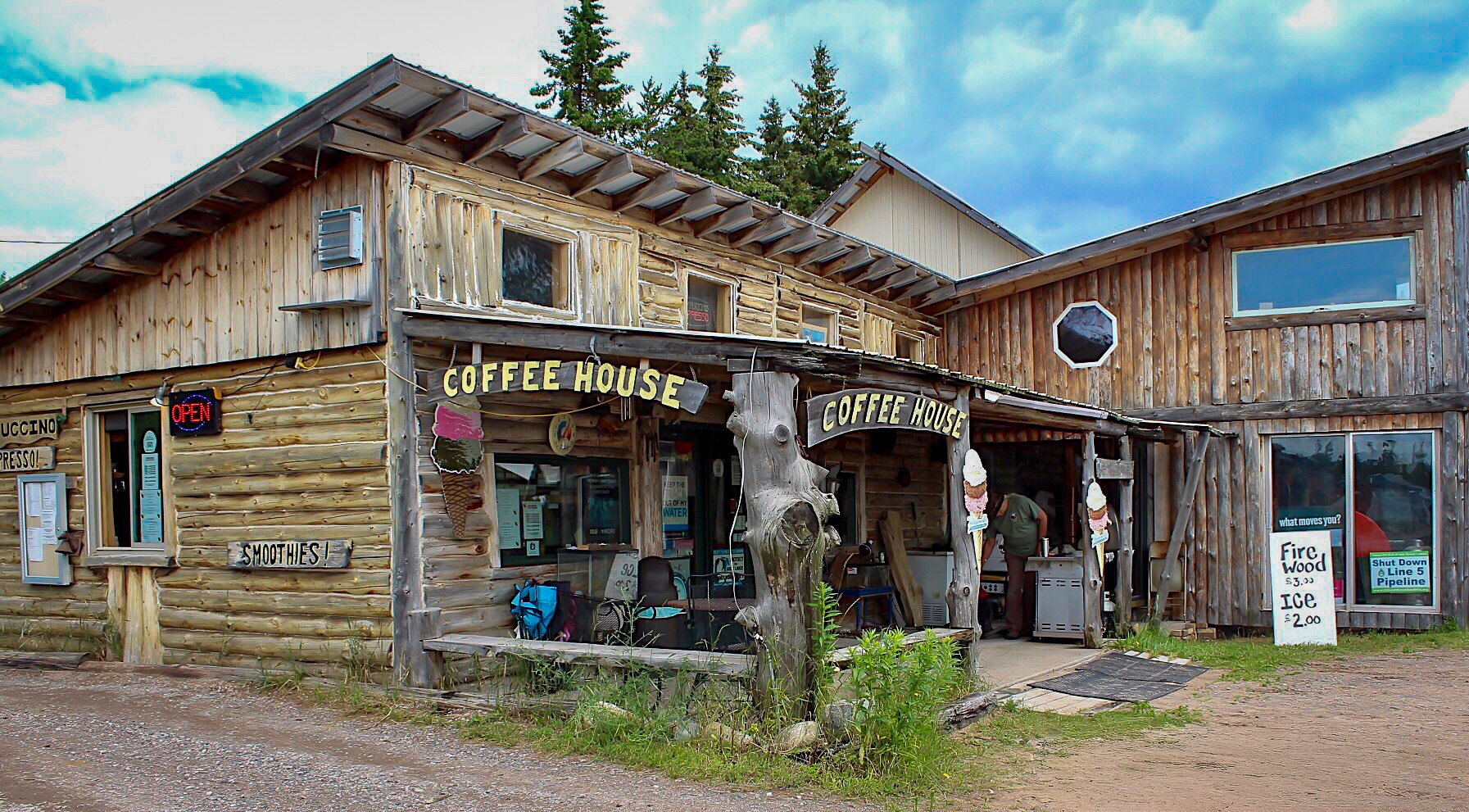 A rustic two-story wooden coffee house with signs advertising coffee, smoothies, ice, and firewood. A glowing "Open" sign is visible, and the building has a shop window, with an outdoor menu and a person working inside. The surroundings are natural w