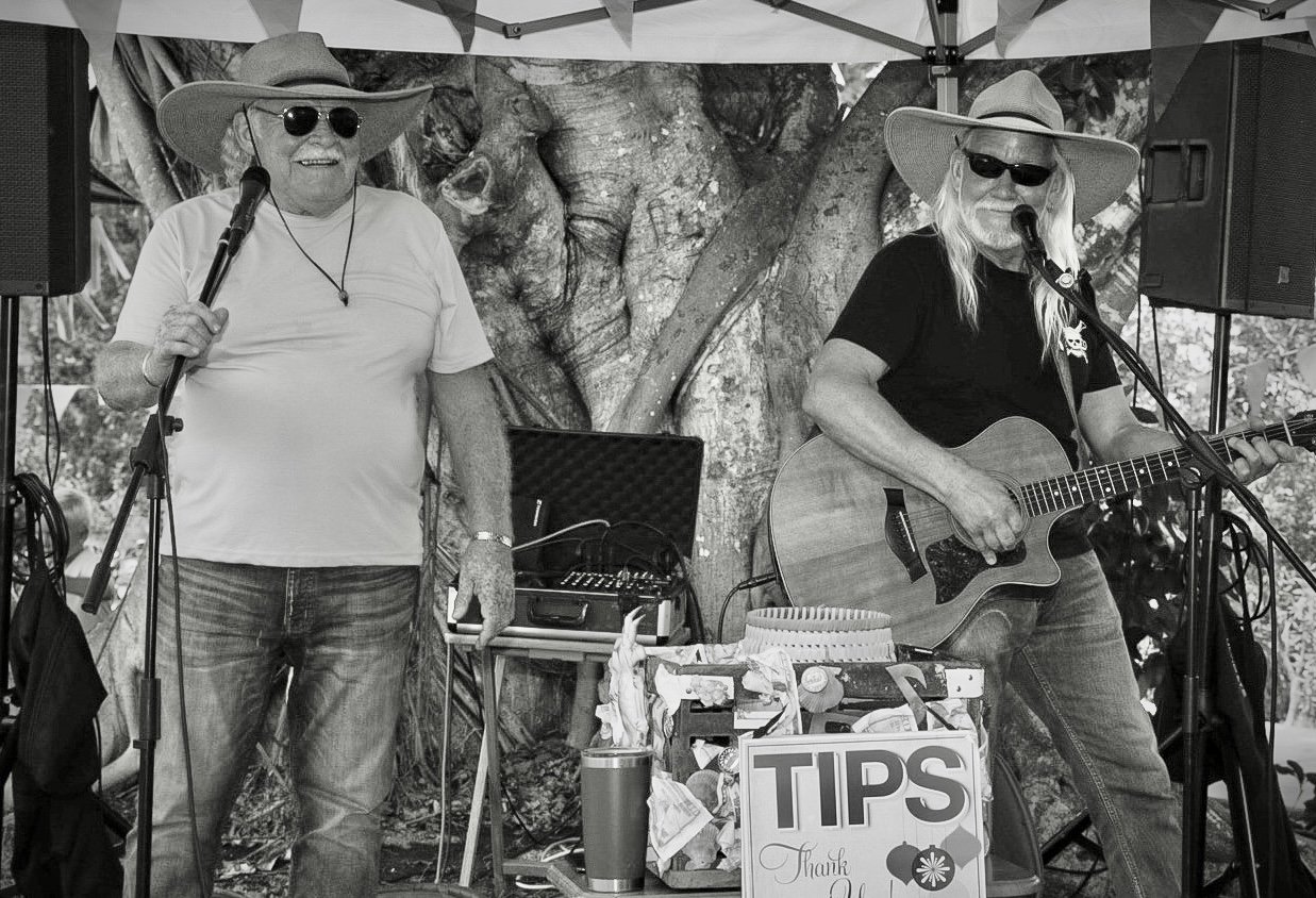 Two elderly men performing music at an outdoor event, one singing into a microphone and the other playing an acoustic guitar, both wearing large floppy hats and sunglasses, with a tree and some equipment in the background.