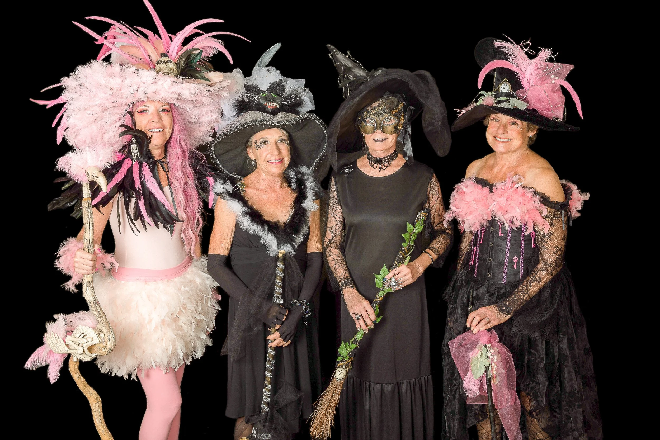 Four women dressed as witches and a witch doctor, wearing elaborate costumes and hats adorned with feathers, skulls, and other decorative elements, standing against a black background.