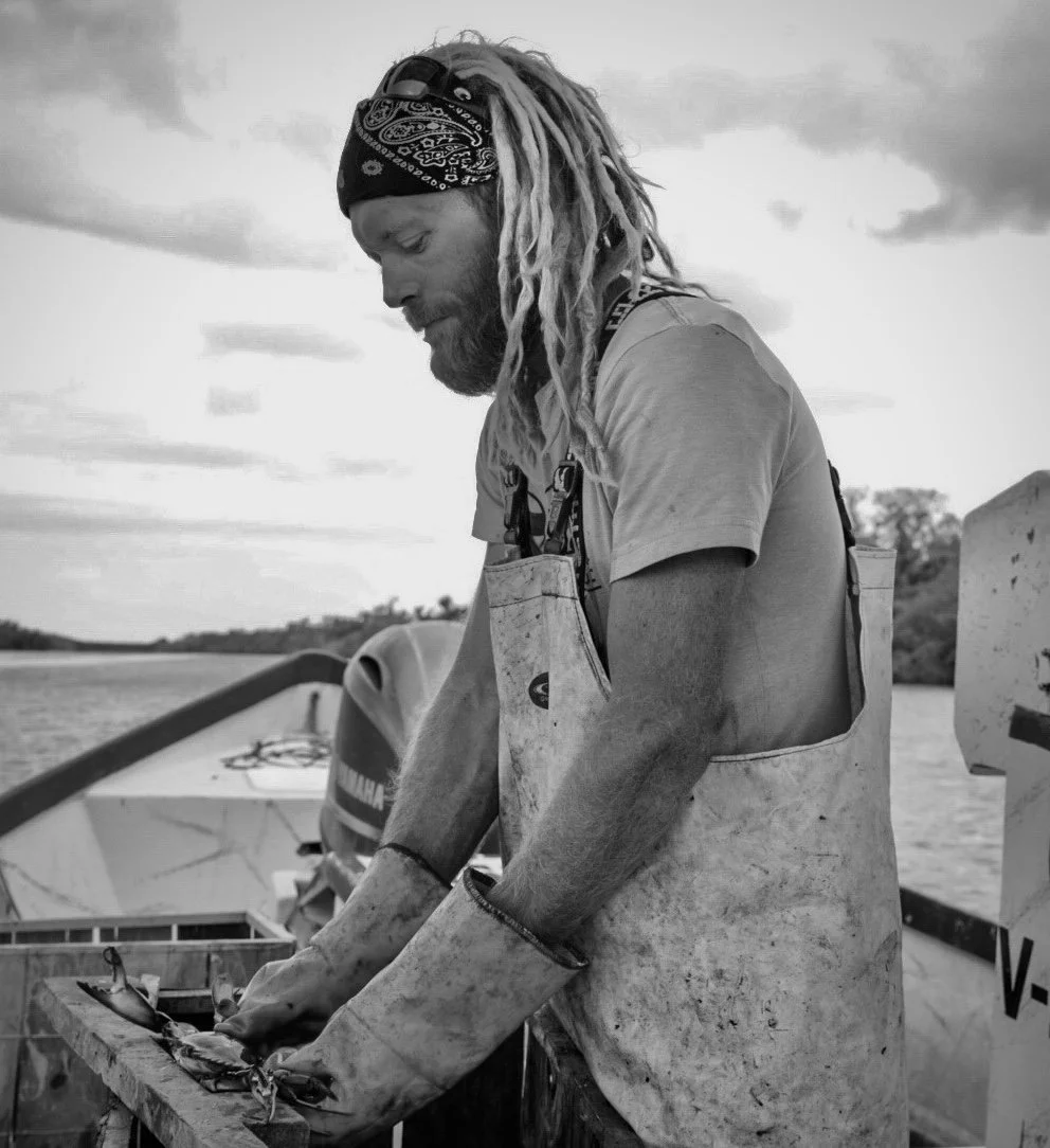 A man with dreadlocks and a bandana working on a boat, handling a fishing net or trap, on a river or lake with a cloudy sky in the background.