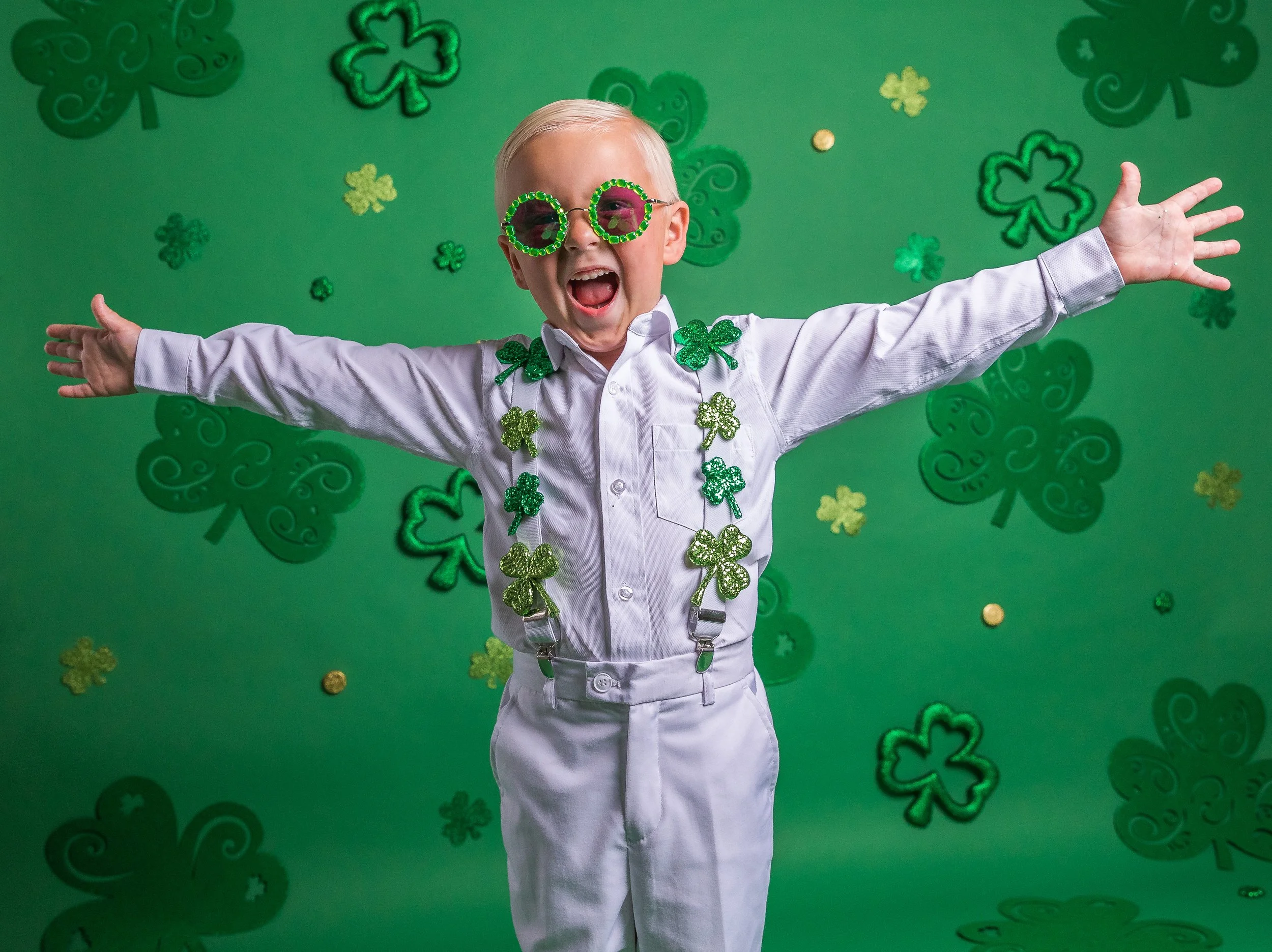 A young boy dressed in white with green glitter shamrock decorations and green shamrock sunglasses in front of a green background decorated with shamrocks and gold coins, celebrating St. Patrick's Day.