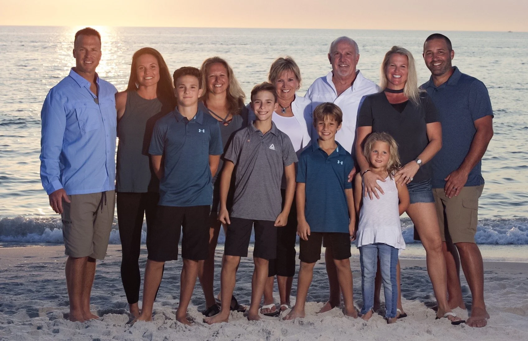 A large family group standing on a sandy beach at sunset, with the ocean and setting sun in the background, smiling at the camera.