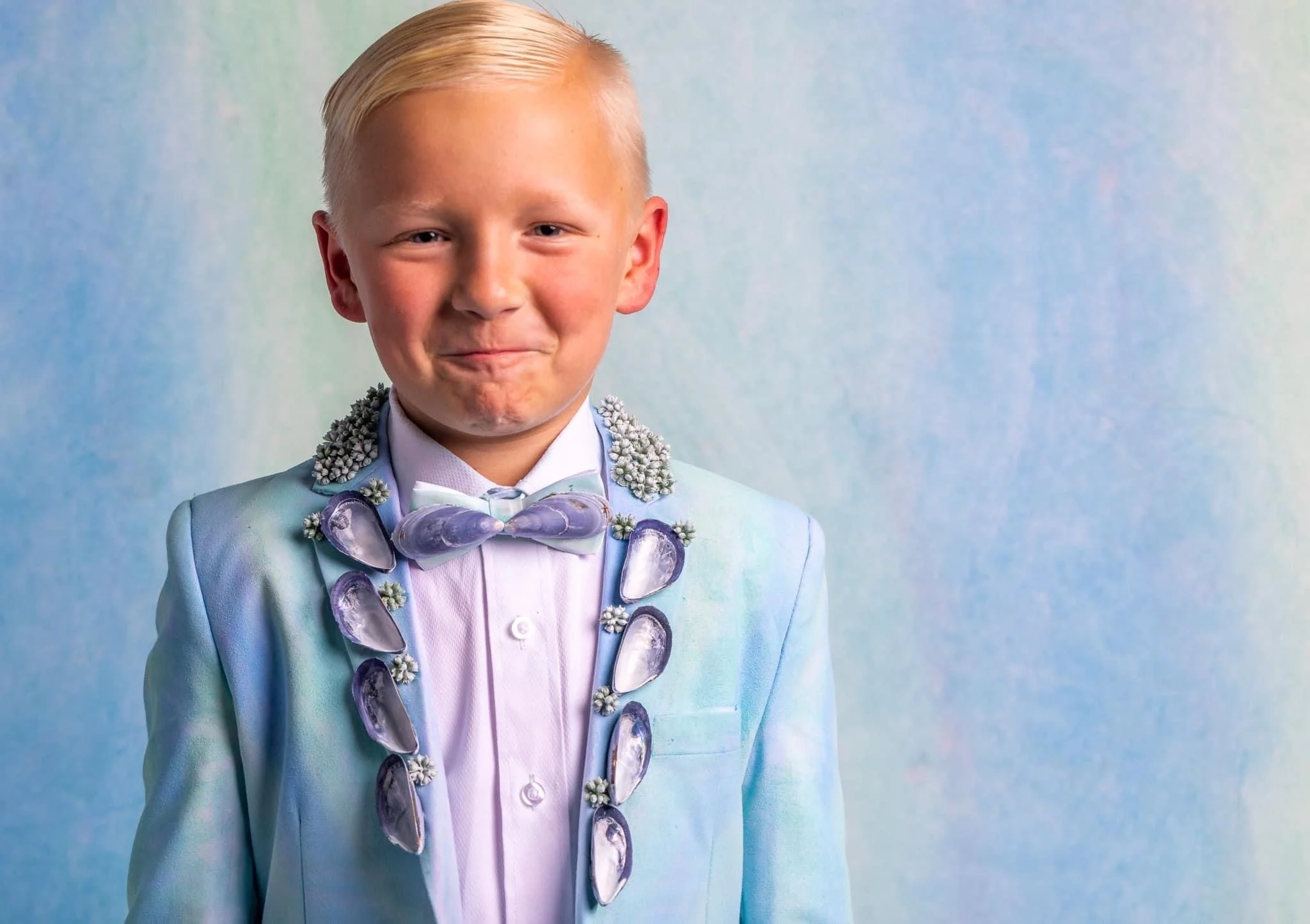 A young boy with blonde hair in a styled haircut, wearing a pastel blue suit with shell and flower decorations, a white shirt, and a lavender bow tie, smiling against a colorful pastel background.