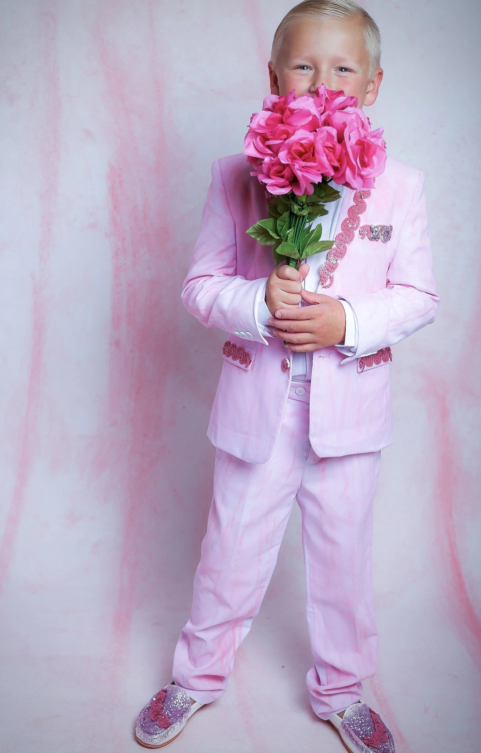 A young boy dressed in a pink suit holding a bouquet of pink flowers and smiling.