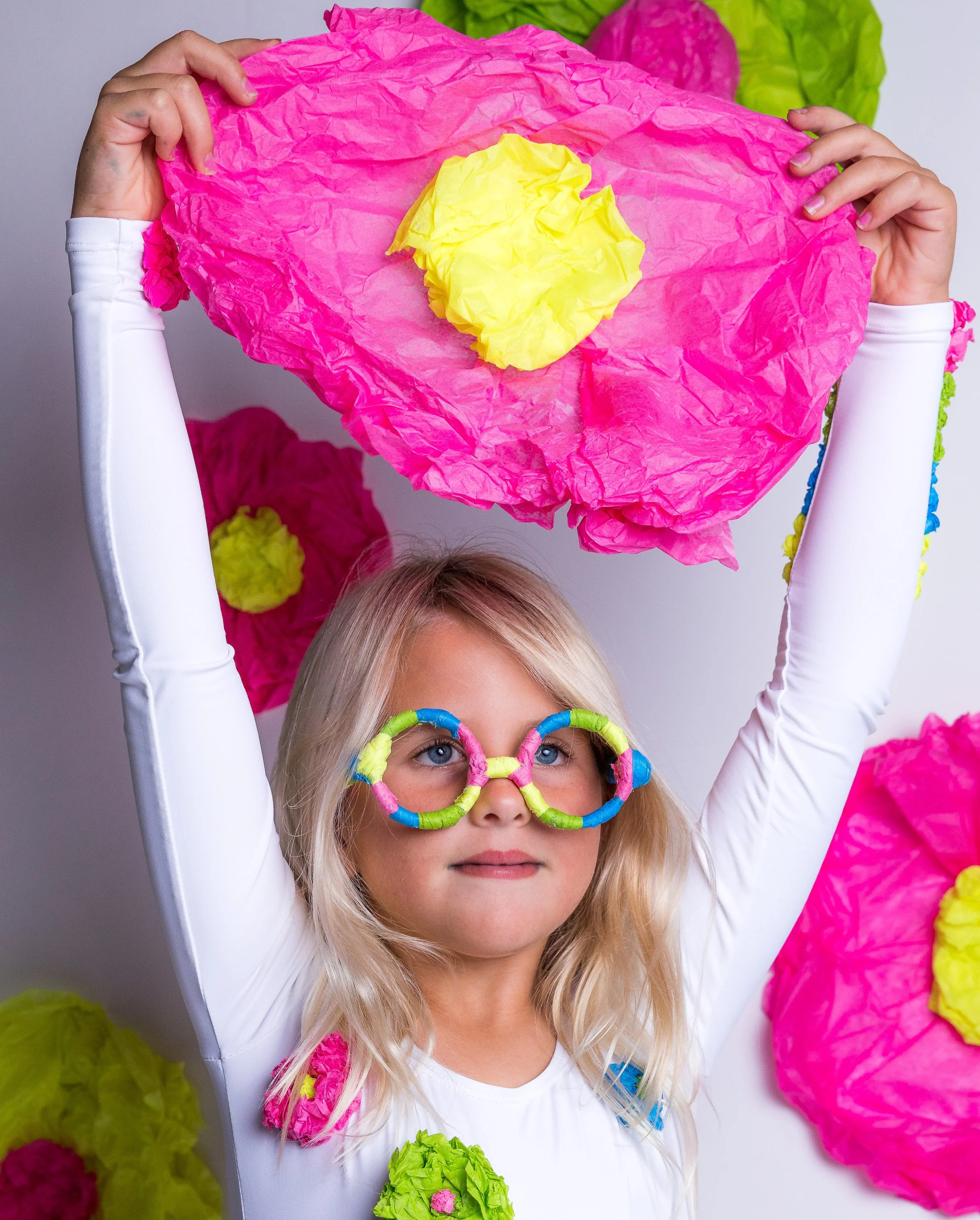 A young girl with blonde hair and blue eyes wearing glasses with colorful frames, holding a large pink tissue paper flower above her head, surrounded by colorful tissue paper flowers on the wall.
