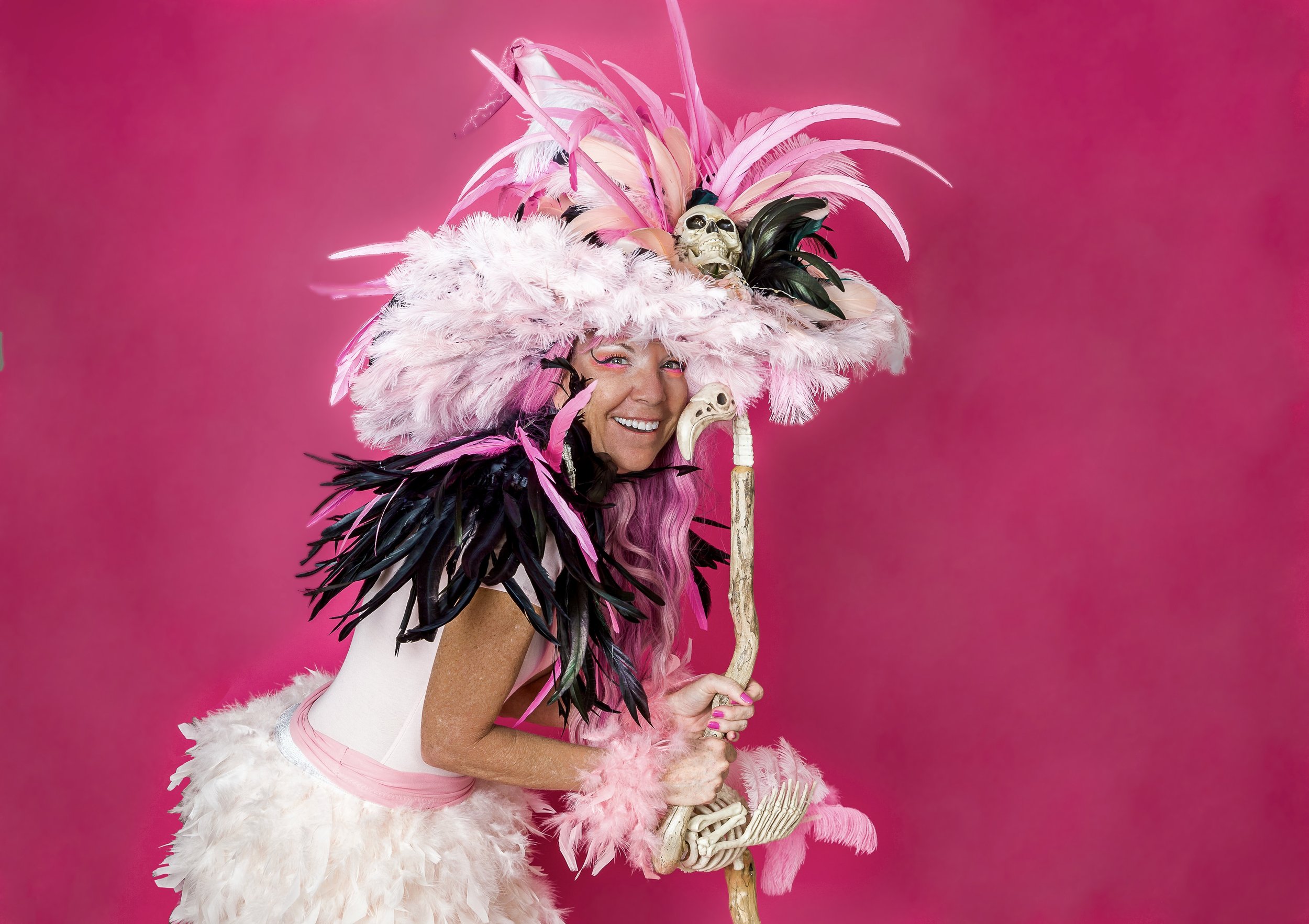  woman wearing a pink feathered headdress with skull decorations, holding a staff with skull and bird elements, dressed in pink and white feathers, standing against a bright pink background.