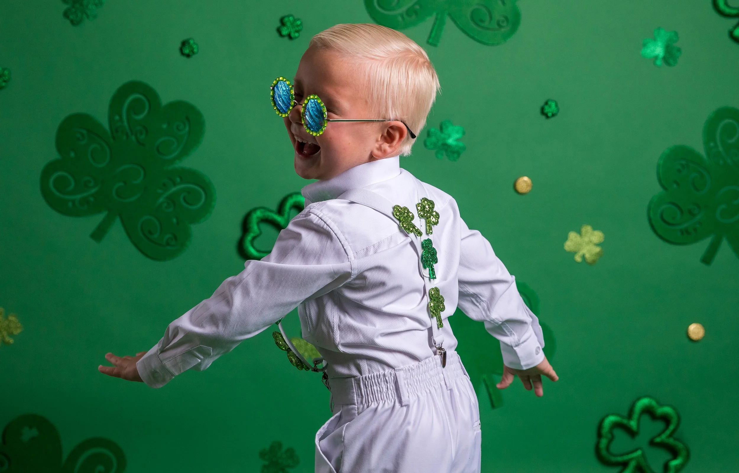 A young child with blonde hair wearing sunglasses decorated with shamrocks, smiling and dancing against a green background with shamrock and gold decorations.