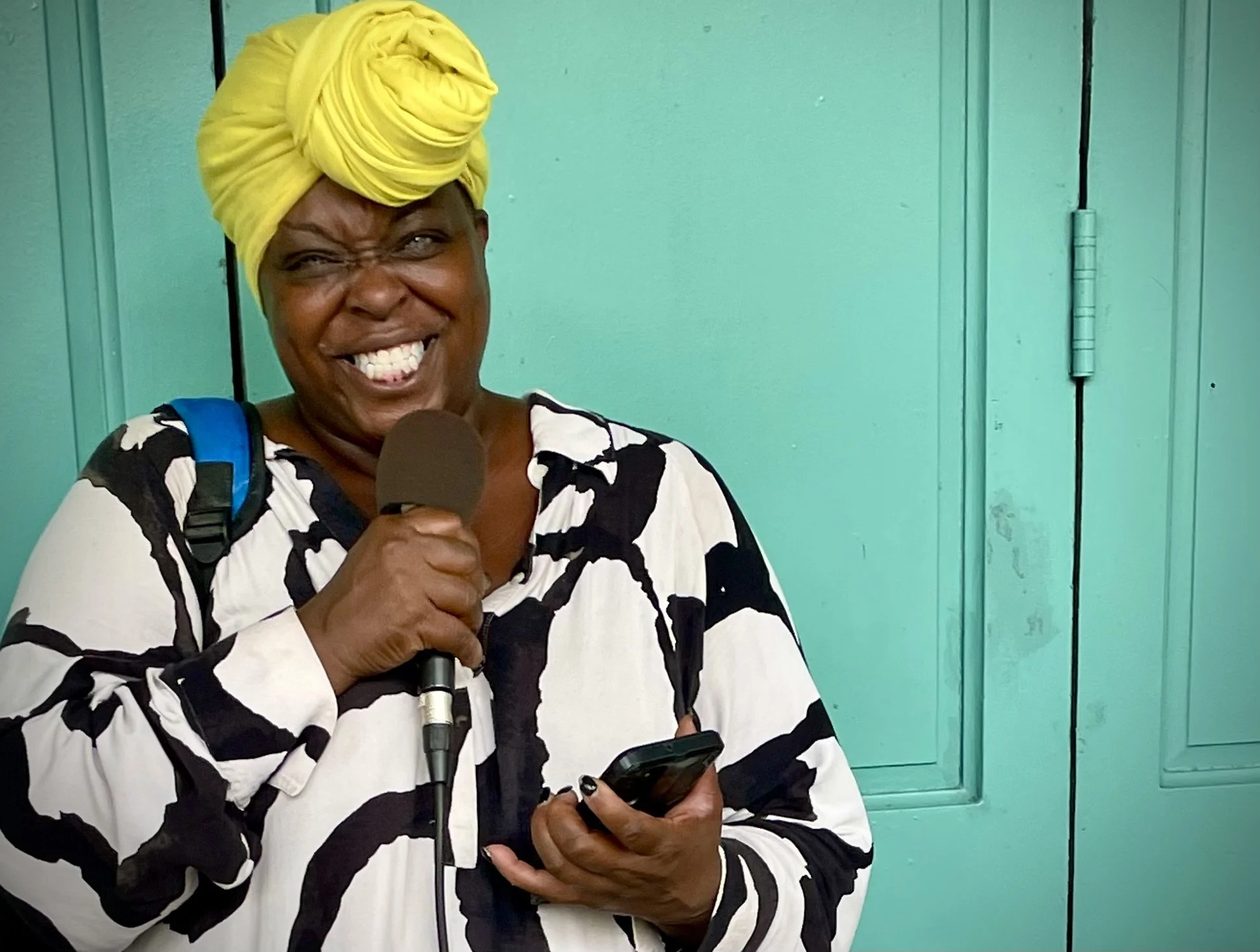 A woman with dark skin wearing a yellow headscarf, black and white patterned top, holding a microphone in one hand and a phone in the other, standing in front of a turquoise-colored vertical wooden slats background.