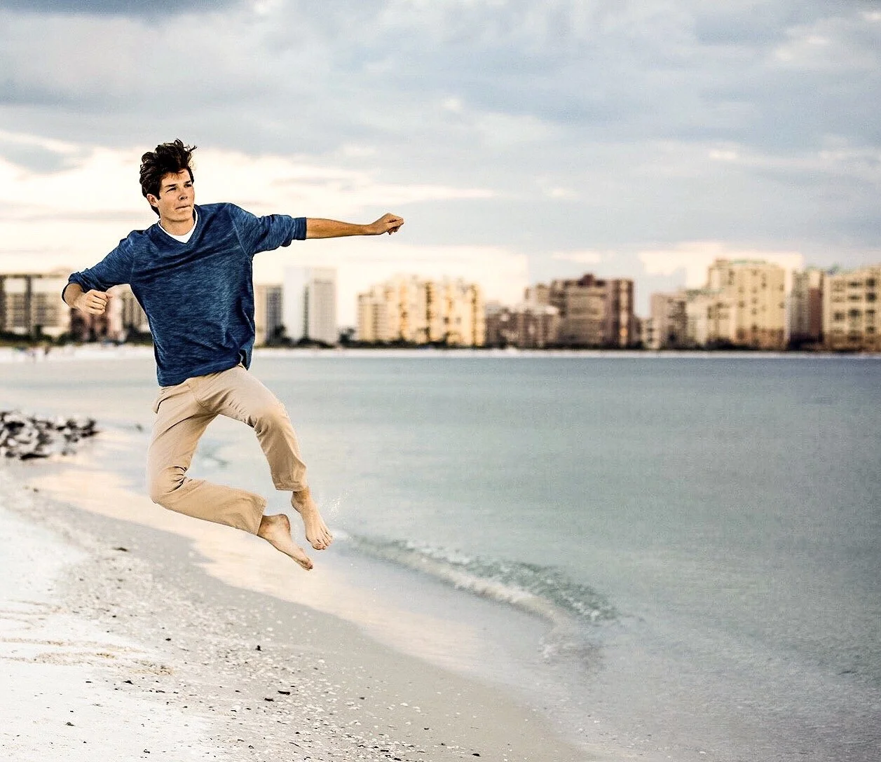 Young man jumping on a sandy beach near water, with city buildings in the background under cloudy sky.