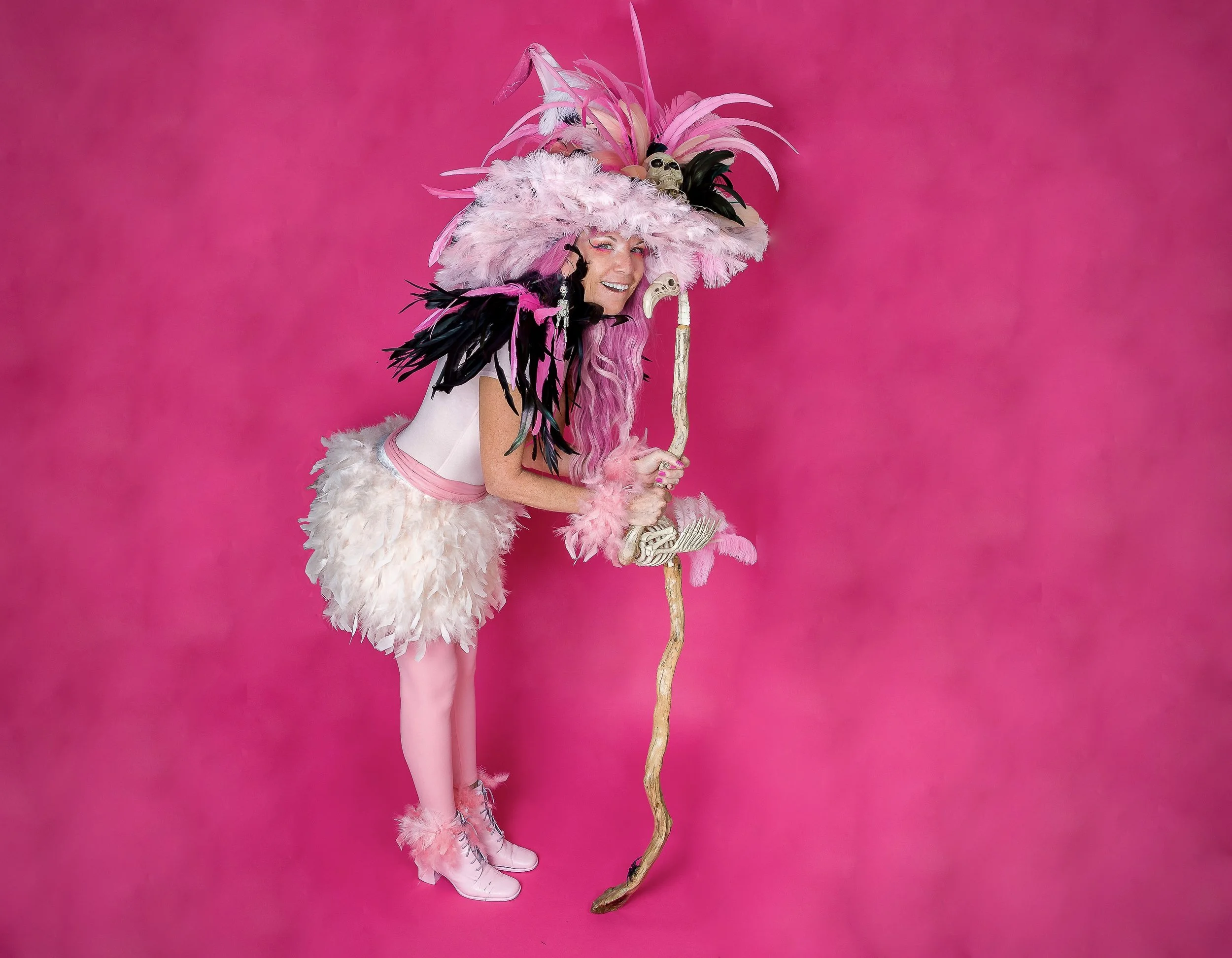 Woman dressed in pink fairy costume with pink feathered hat, feathered wings, pink tights, and pink shoes, holding a staff with skulls and feathers, standing against a pink background.
